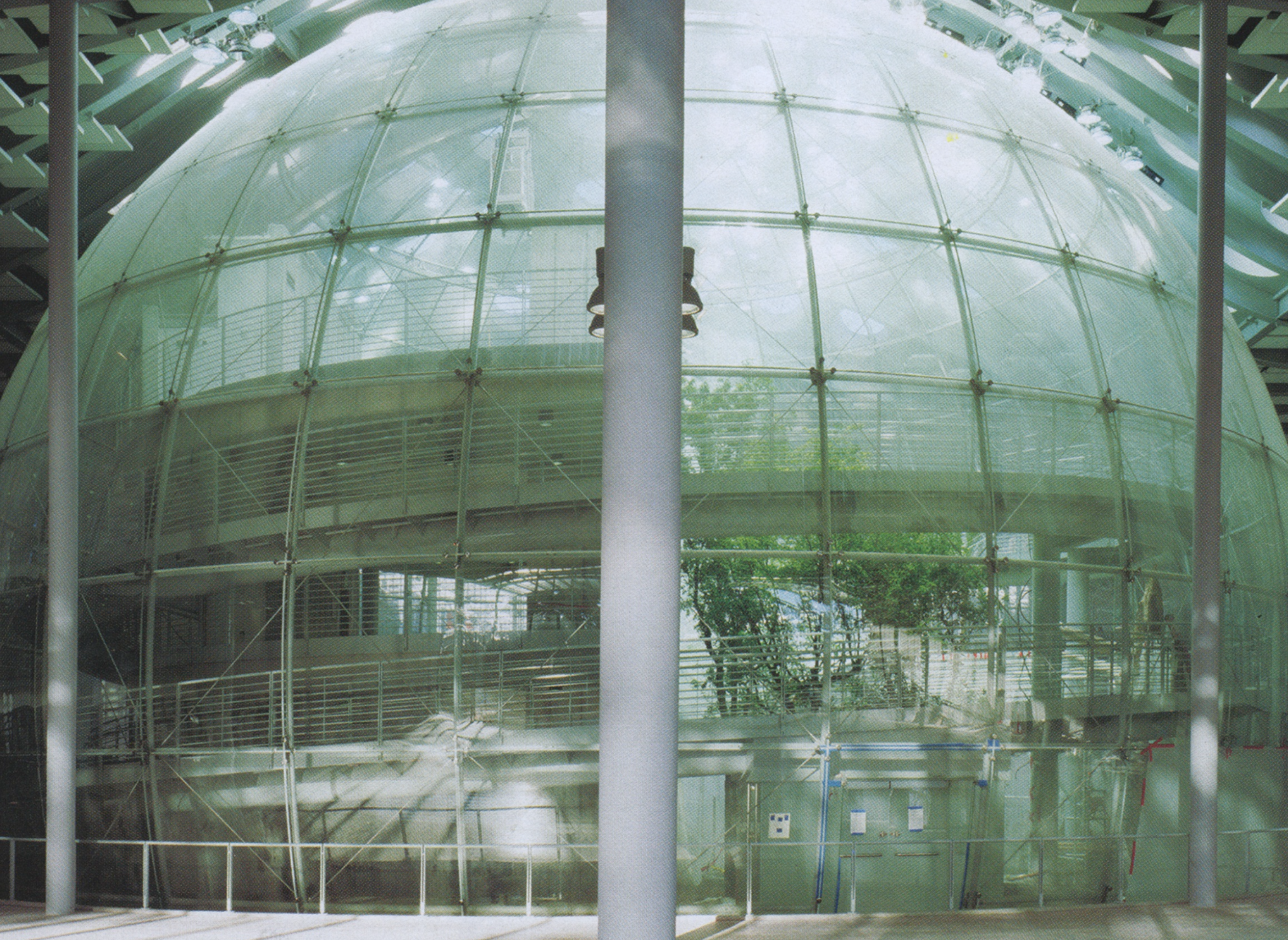 Contemporary building with large curved glass facade, supported by metal framing and vertical pillars, with visible interior stairs and greenery outside.