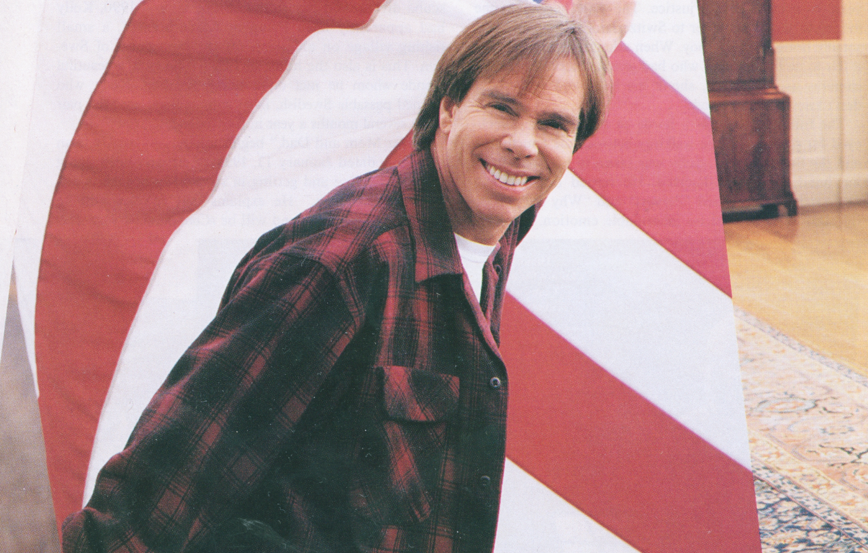 A smiling man with light brown hair wearing a red plaid shirt. He is positioned in front of a large American flag with red, white, and blue stars, possibly at a celebration or event.