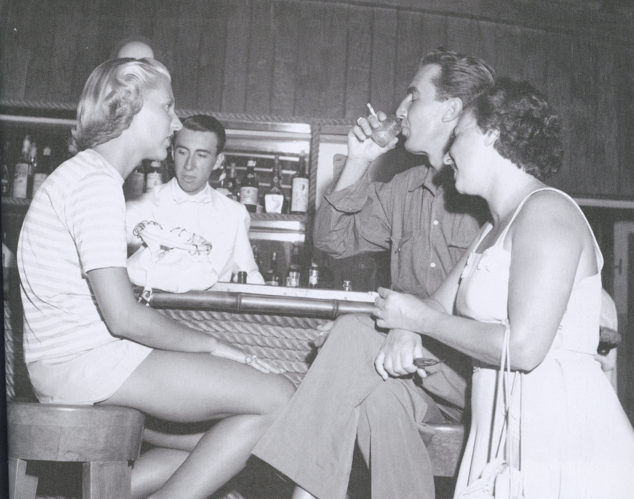 A black and white photo of four people in a bar. Two women and two men are sitting and standing, engaged in conversation. One woman is holding a cigarette, and the other has her arm resting on her knee. A man is drinking from a glass, and the other man is standing behind the bar.