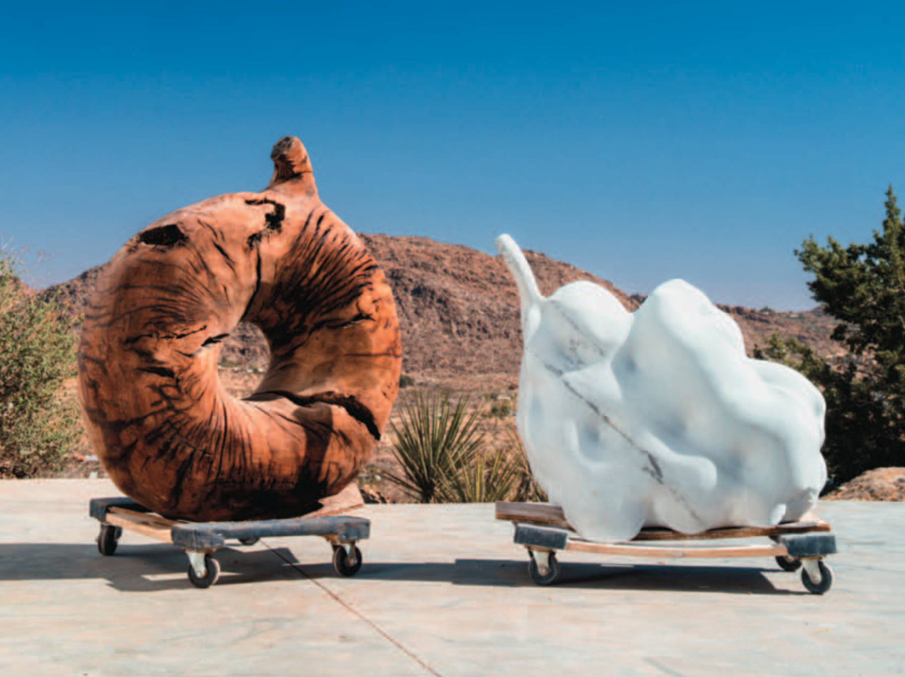 Two sculptures on wheeled platforms in a desert landscape with mountains and shrubs, one sculpture resembling a brown animal and the other a white abstract shape.