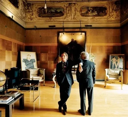 Two men in suits standing in an art gallery or museum, looking at artwork. The room has wooden paneling, a framed portrait behind them, and artwork on either side. There are chairs, tables, and decorative paintings on the walls and ceiling.