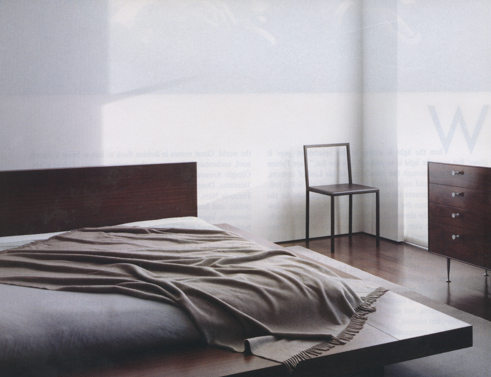 A minimalist bedroom with a bed that has a dark wooden headboard and a beige blanket, a black chair near the corner, and a dark wooden dresser with four drawers. The room has white walls and a hardwood floor, with sunlight coming through the windows.