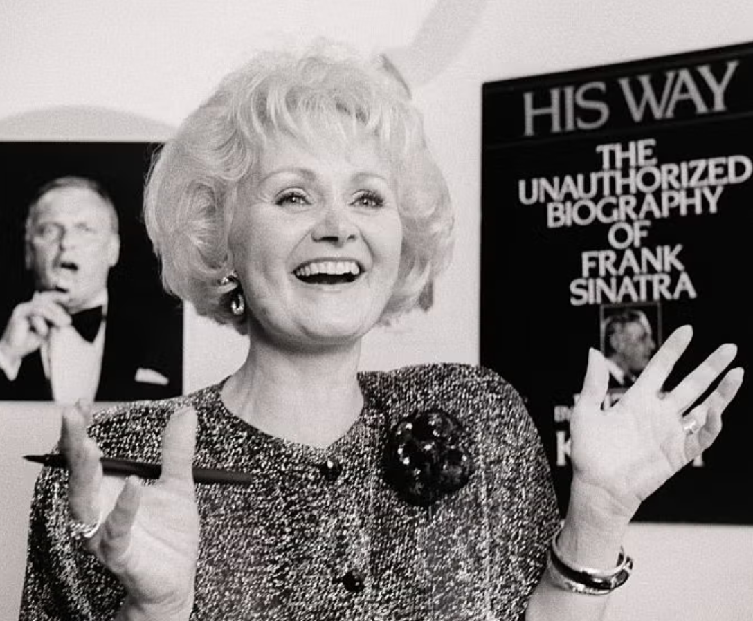 Black and white photo of a cheerful woman with curly hair, holding a book and smiling, with a poster of Frank Sinatra in the background.