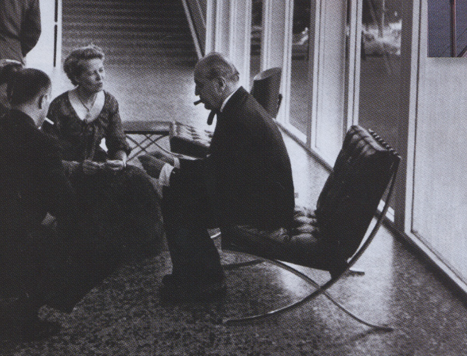 A man with a pipe sitting on a modern rocking chair near three children sitting on the floor, inside a room with large windows.