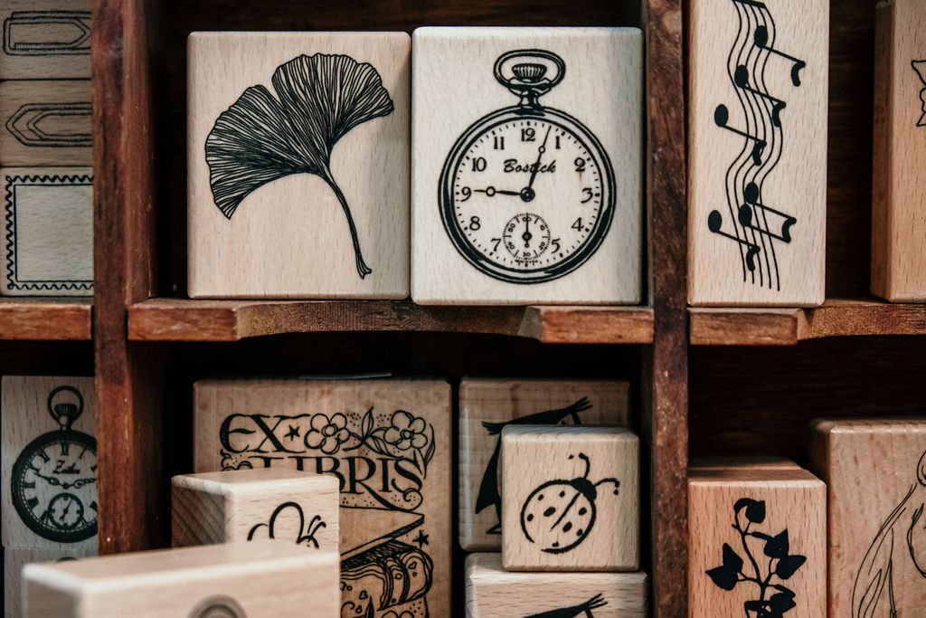 Wooden blocks with black illustrations including a ginkgo leaf, a stopwatch, musical notes, a ladybug, a flower with the word 'EX LIBRIS,' and a sunflower.