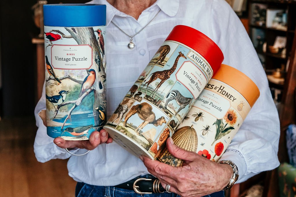 Person holding three cylindrical vintage puzzle tins with bird, animal, and floral designs.
