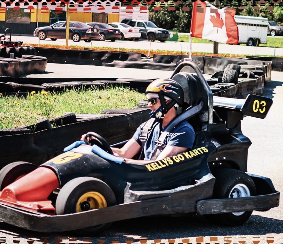 A person driving a go-kart on a race track at Kelly's Go Karts, wearing a helmet and sunglasses. A Canadian flag is visible in the background. The go-kart is black with yellow number 03.