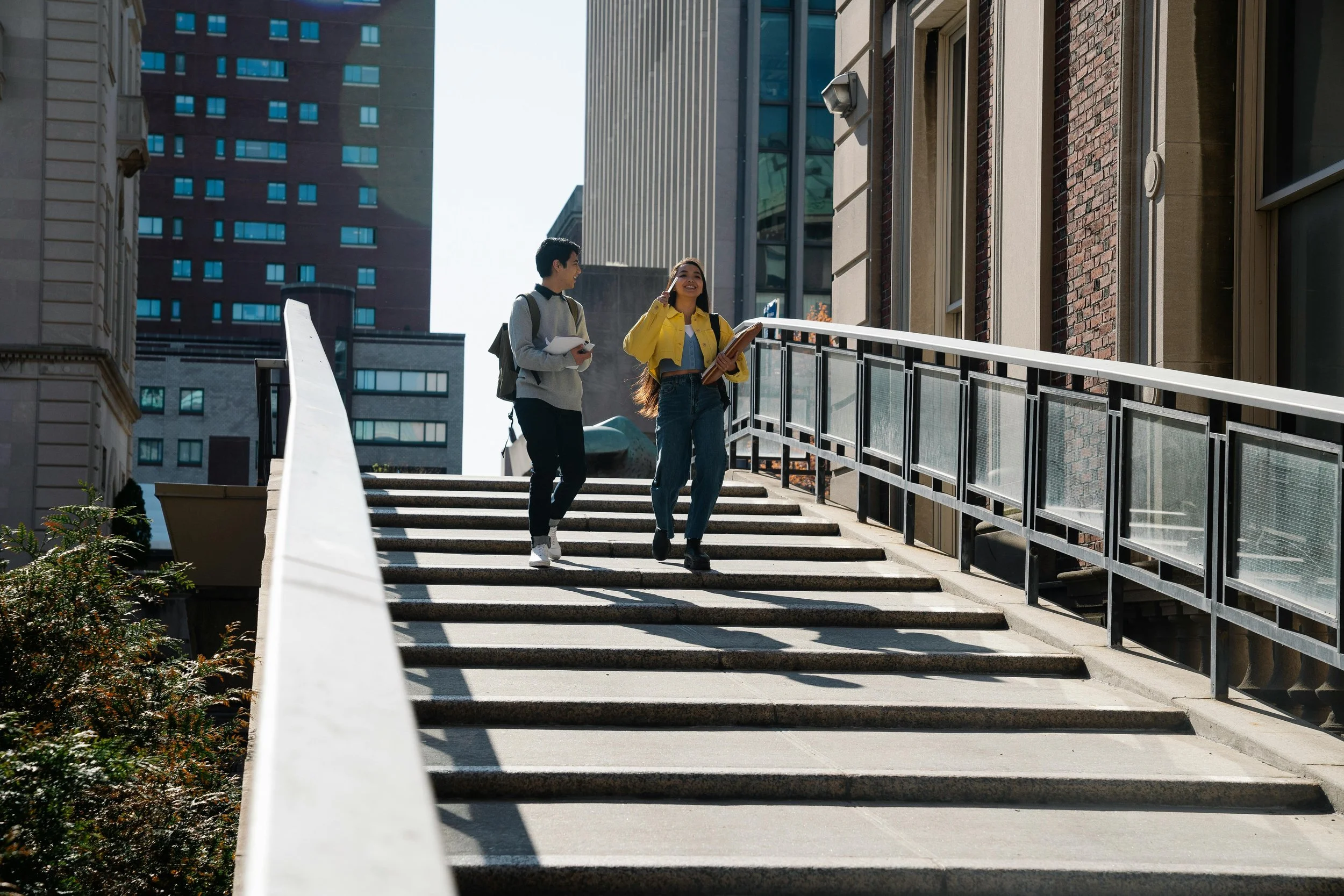 Two people walking down outdoor steps near tall buildings, one holding a book and wearing a yellow jacket, the other carrying a backpack.
