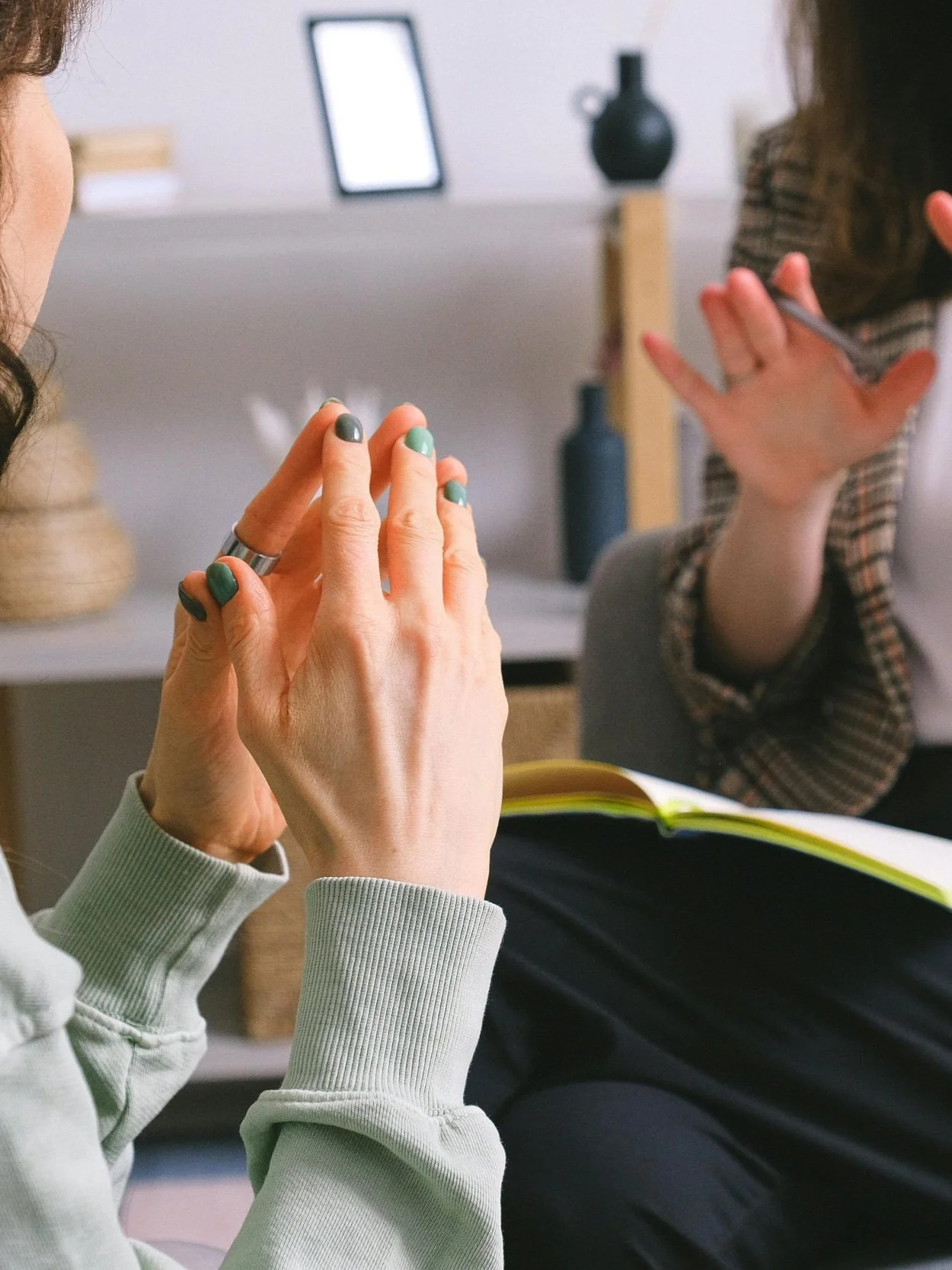 Close-up of two people in a meeting, one with green nail polish gesturing, the other holding a pen and wearing a plaid jacket. A notebook is visible on the lap of one person.