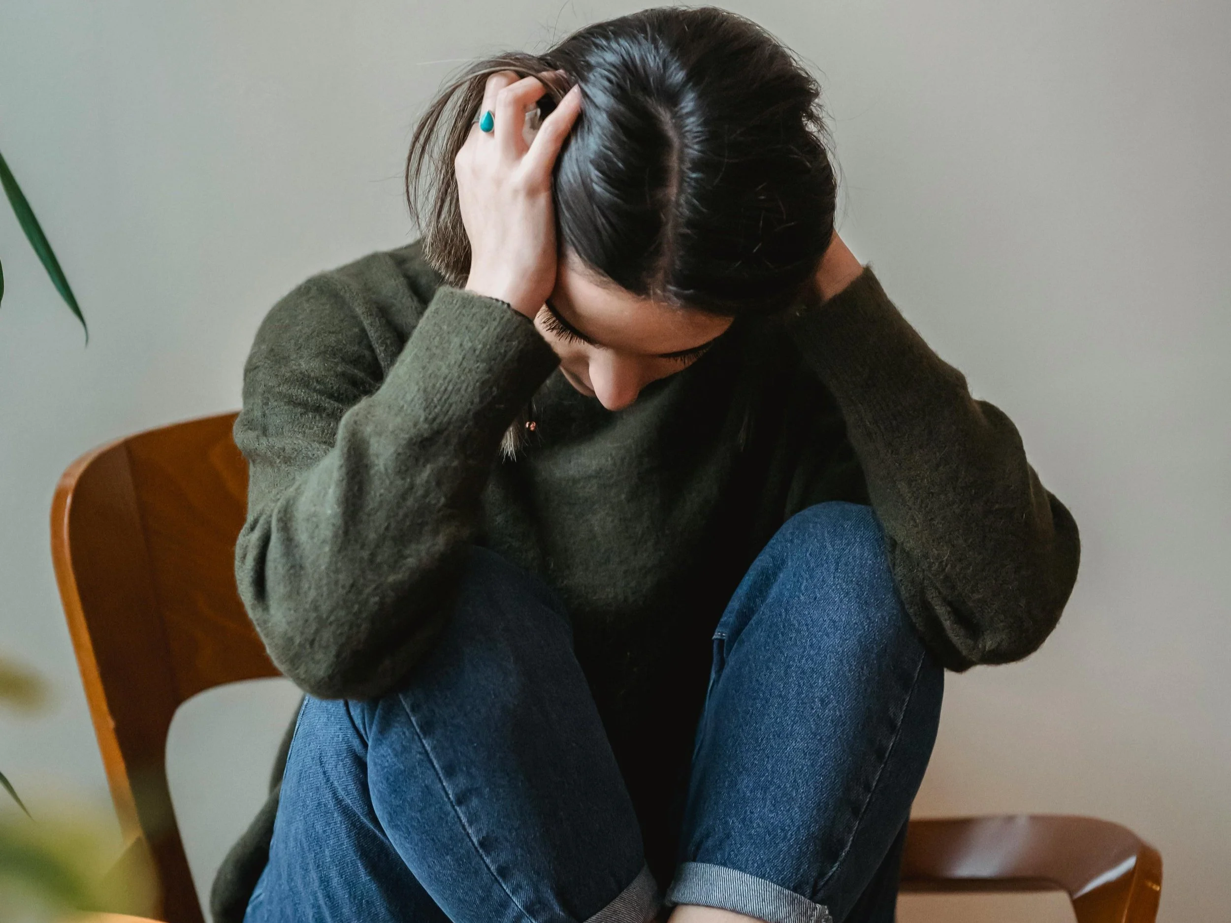 A woman sitting on a wooden chair with her head bowed and hands clutching her head, appearing distressed or upset.
