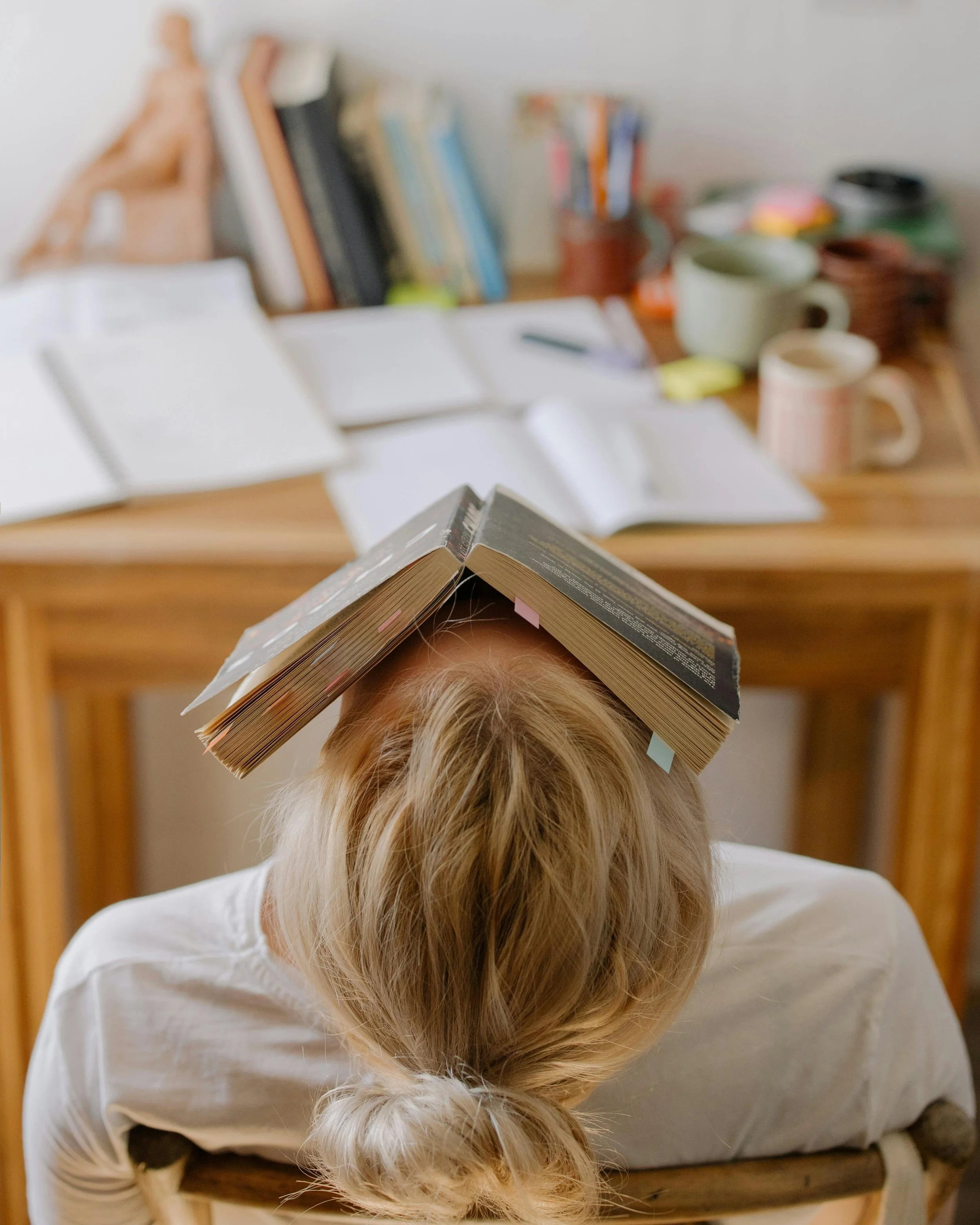 Person with blonde hair sitting at a desk with an open book on their head, surrounded by open notebooks, books, mugs, and stationery on the desk in a cluttered workspace.