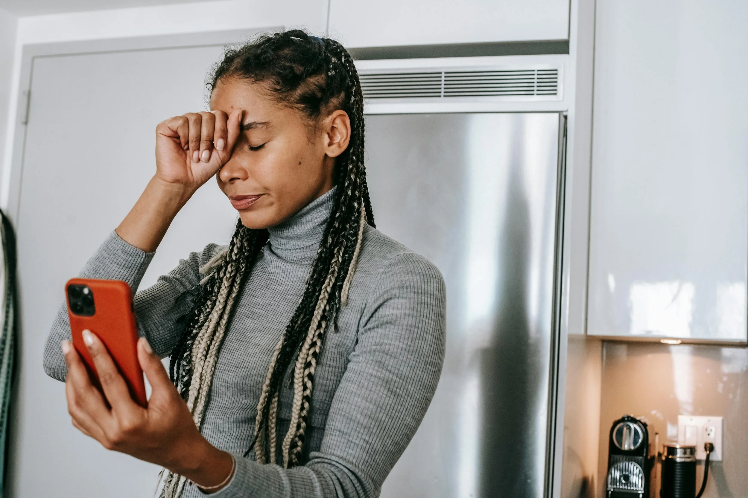 A woman with braids and a gray turtleneck holding a red smartphone, appearing upset or stressed, in a kitchen.