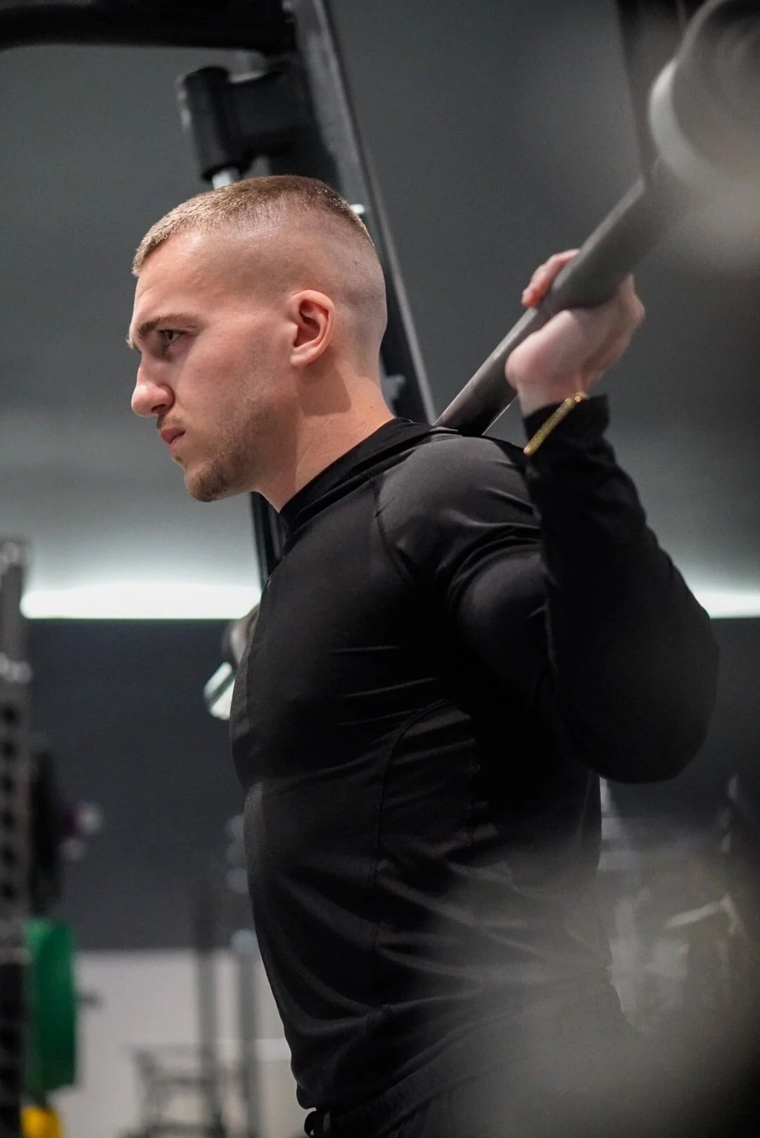 Un homme musclé, avec une coupe de cheveux courte, soulève une barre d'haltères lors d'une séance d'entraînement en salle de sport, portant un maillot ou t-shirt noir.