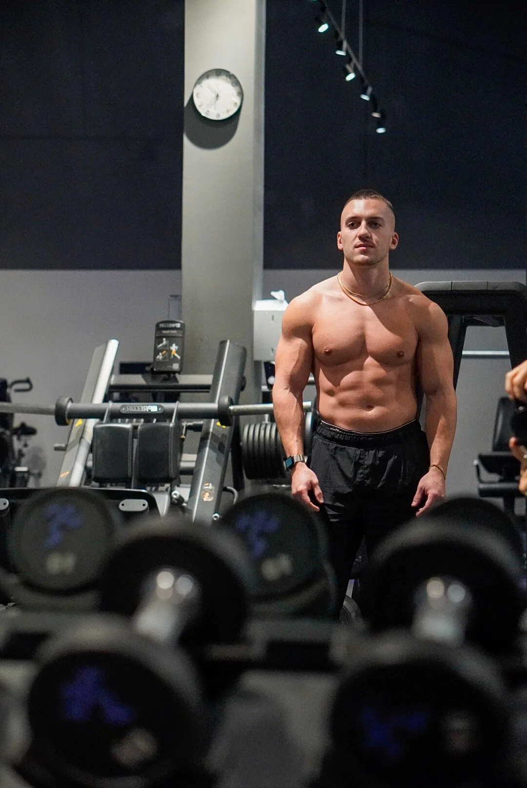 Un homme musclé sans chemise dans une salle de sport, entouré d'haltères et d'équipements d'exercice, avec une montre et un collier, sous un éclairage tendance.