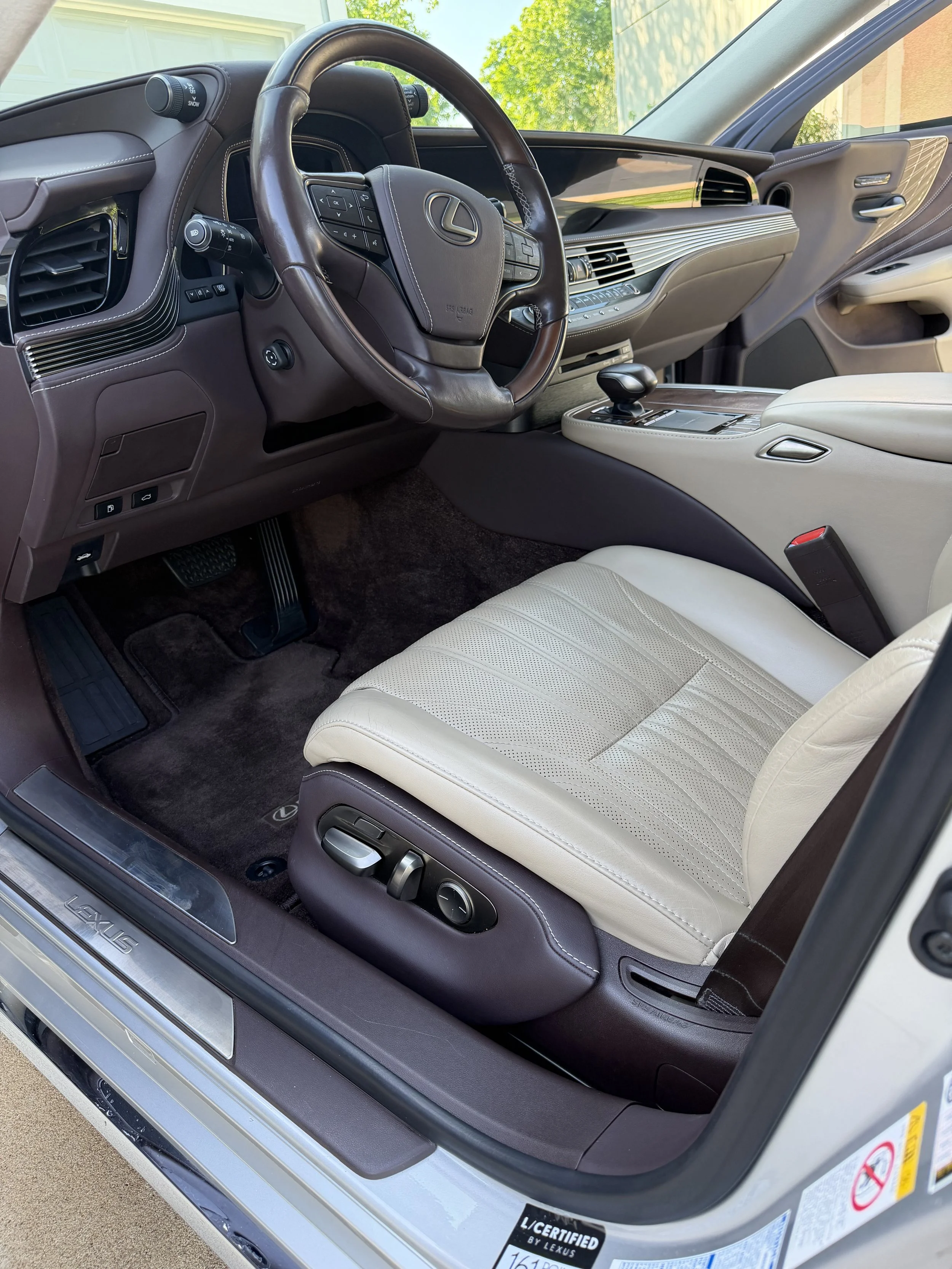 Interior of a Lexus vehicle, featuring a beige leather seat, a brown dashboard with a steering wheel, gear shifter, and various controls, with the door open showing the Lexus badge and a sticker indicating the vehicle's certification.