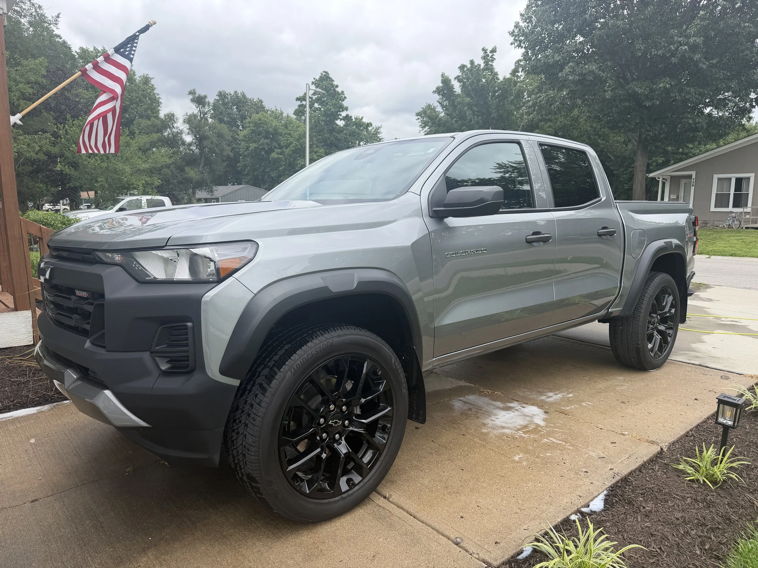 A silver Chevrolet Colorado pickup truck parked on a driveway with a American flag on a wooden pole in the background, greenery, and a grey house.