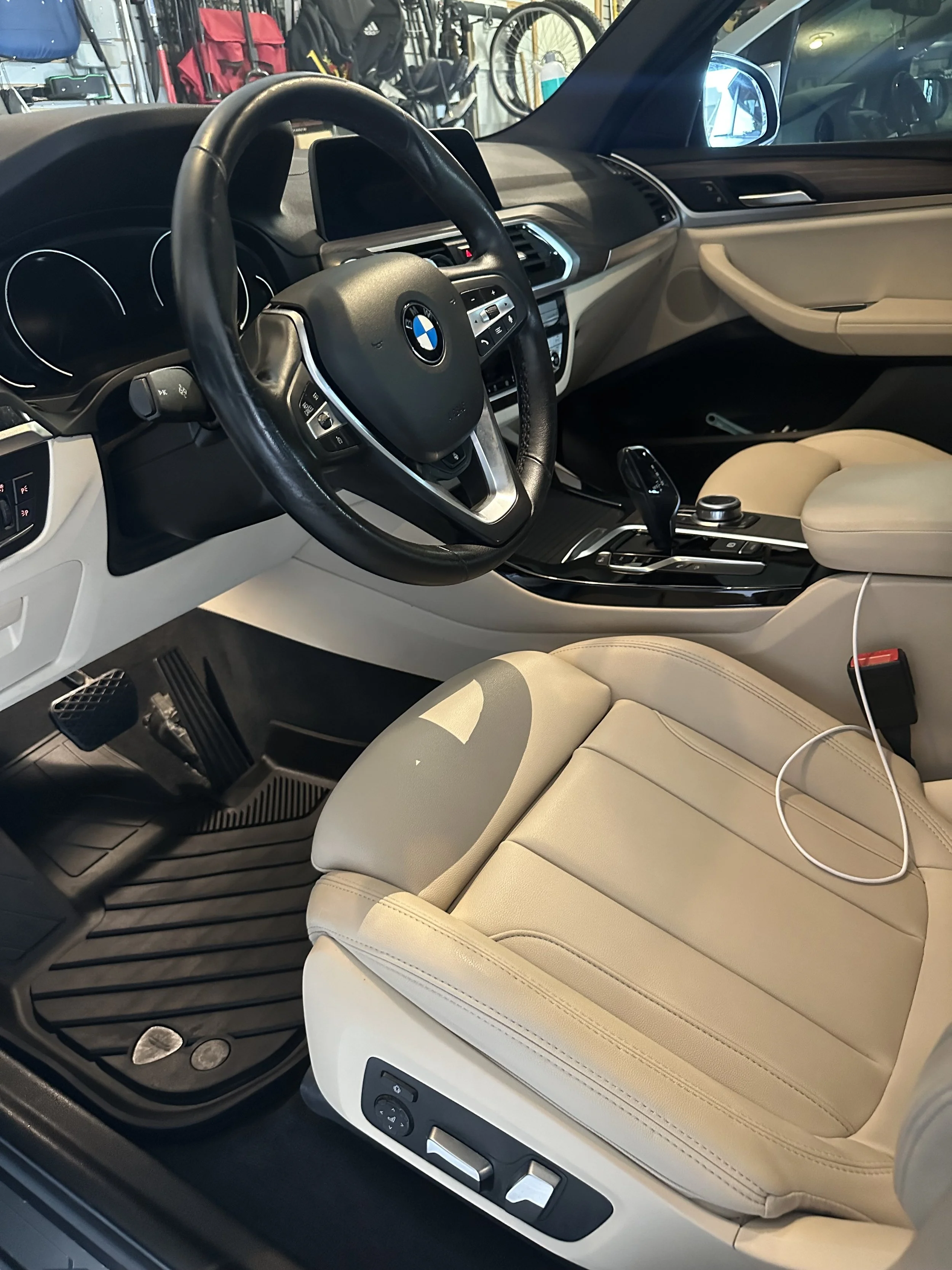 Interior of a BMW car showing the steering wheel, beige leather seats, gear shift, and dashboard.