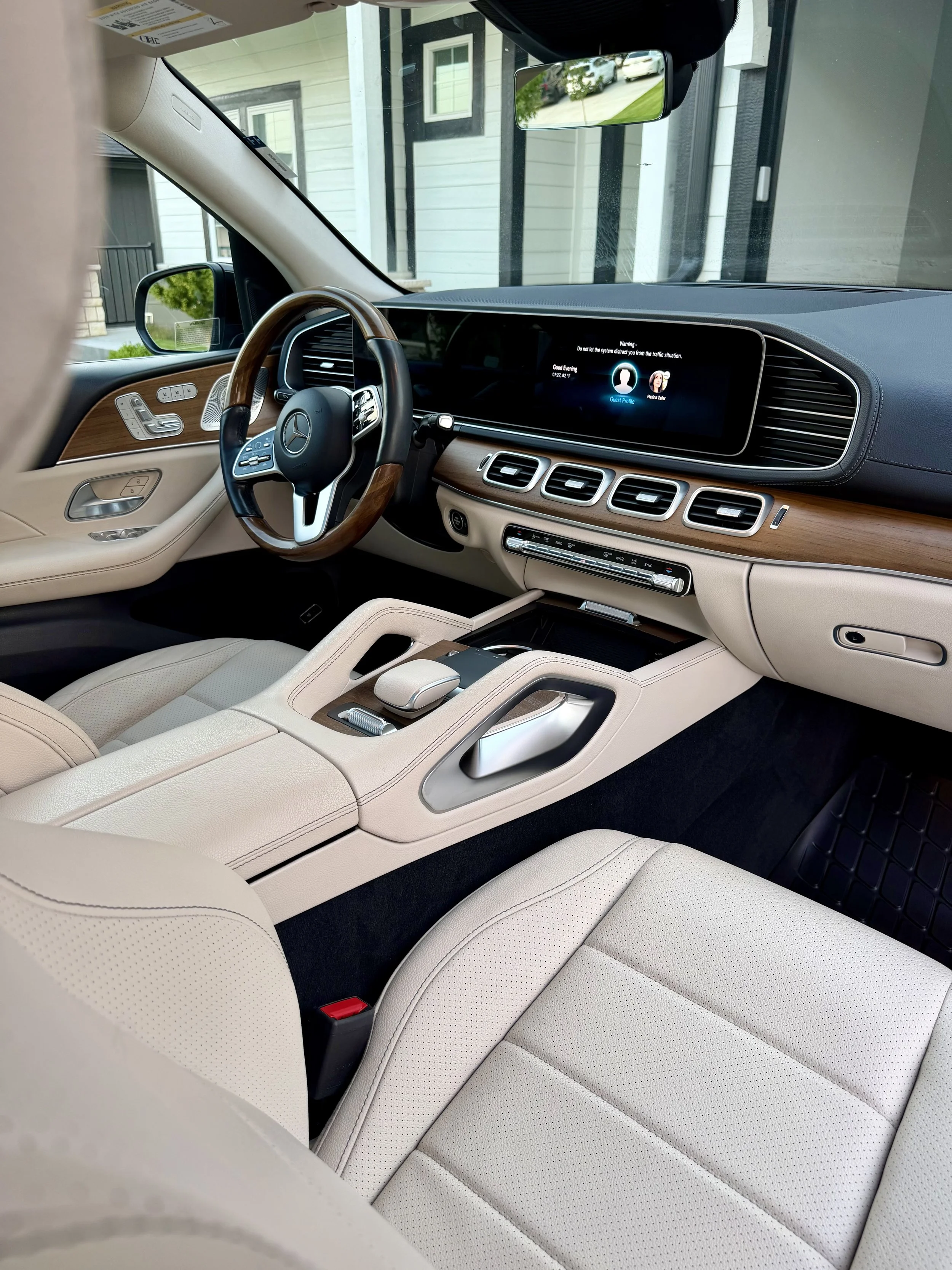 The interior of a luxury Mercedes-Benz vehicle with a cream-colored leather seat, wooden trim, and a digital dashboard displaying a guest profile, parked in a driveway with a house visible through the windows.