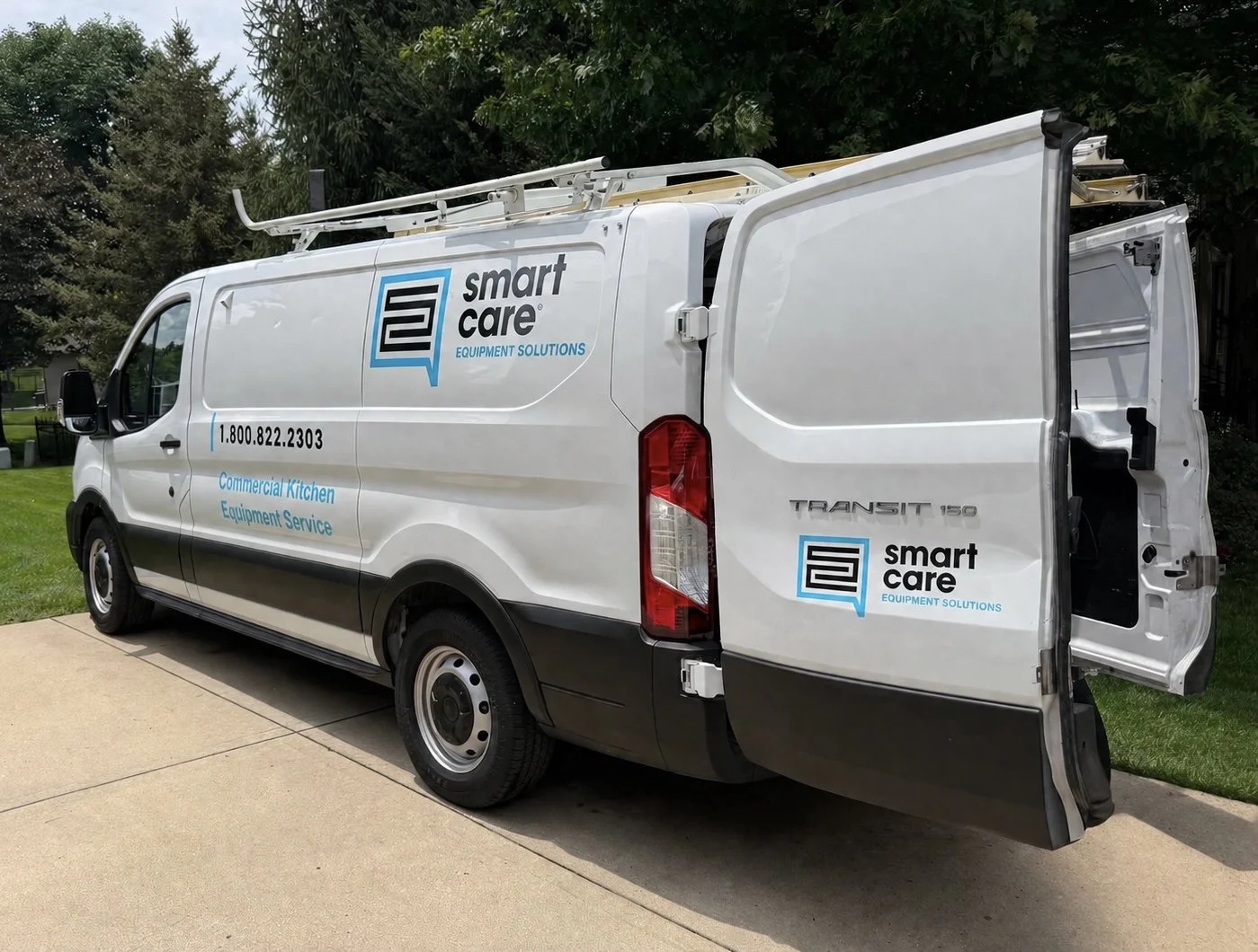 White commercial van with Smart Care logo and contact information, parked outside on a driveway, surrounded by green trees and grass.