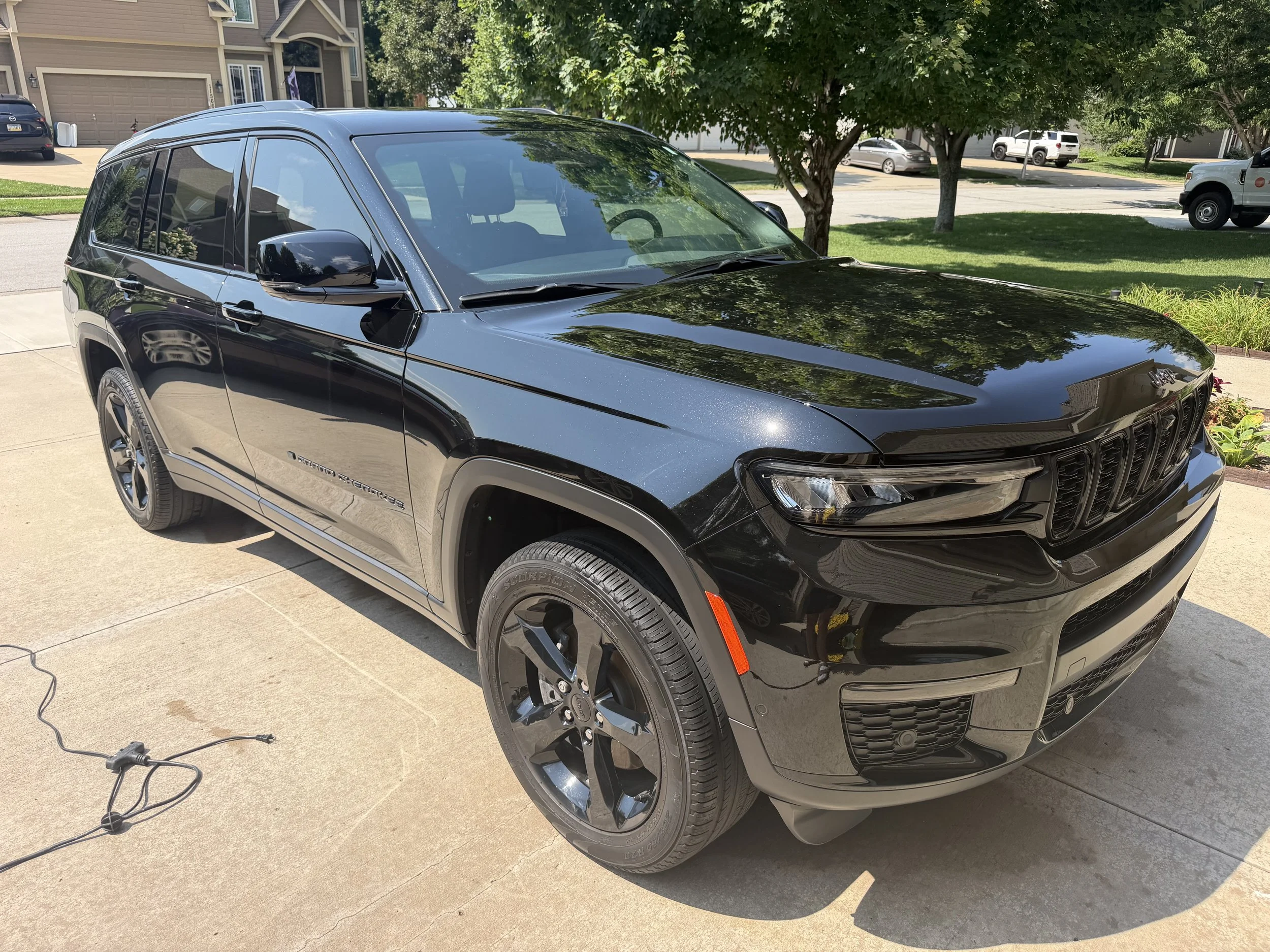 A black Jeep Grand Cherokee parked on a driveway in a suburban neighborhood with trees and other cars in the background.