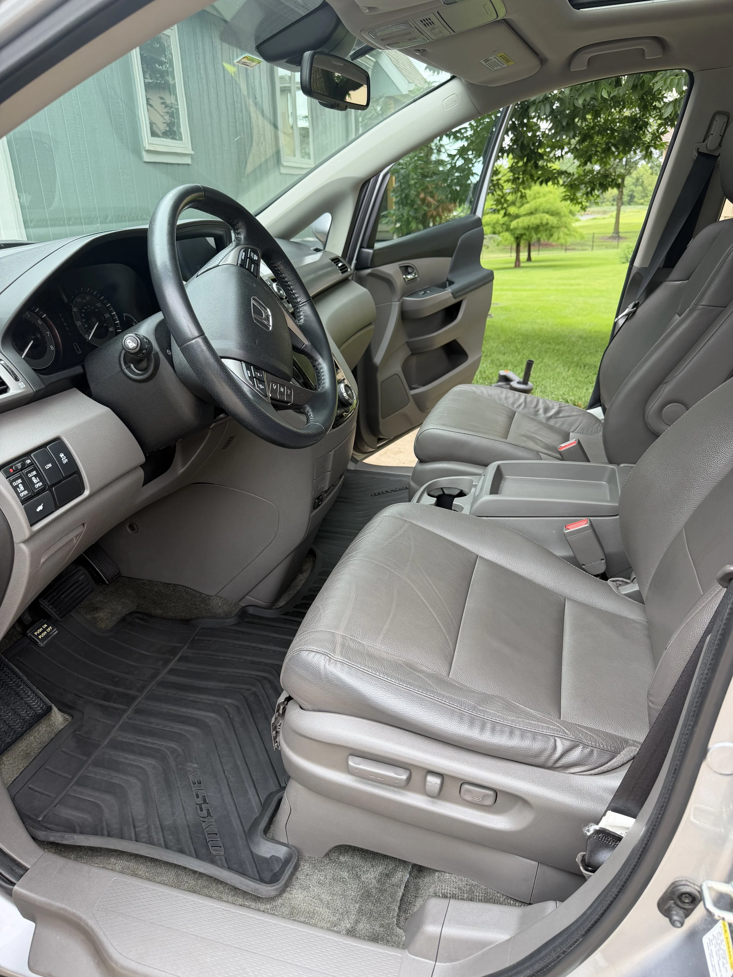 Interior of a Honda vehicle showing the front driver and passenger seats, steering wheel, dashboard, and windows overlooking a green outdoor area with trees.