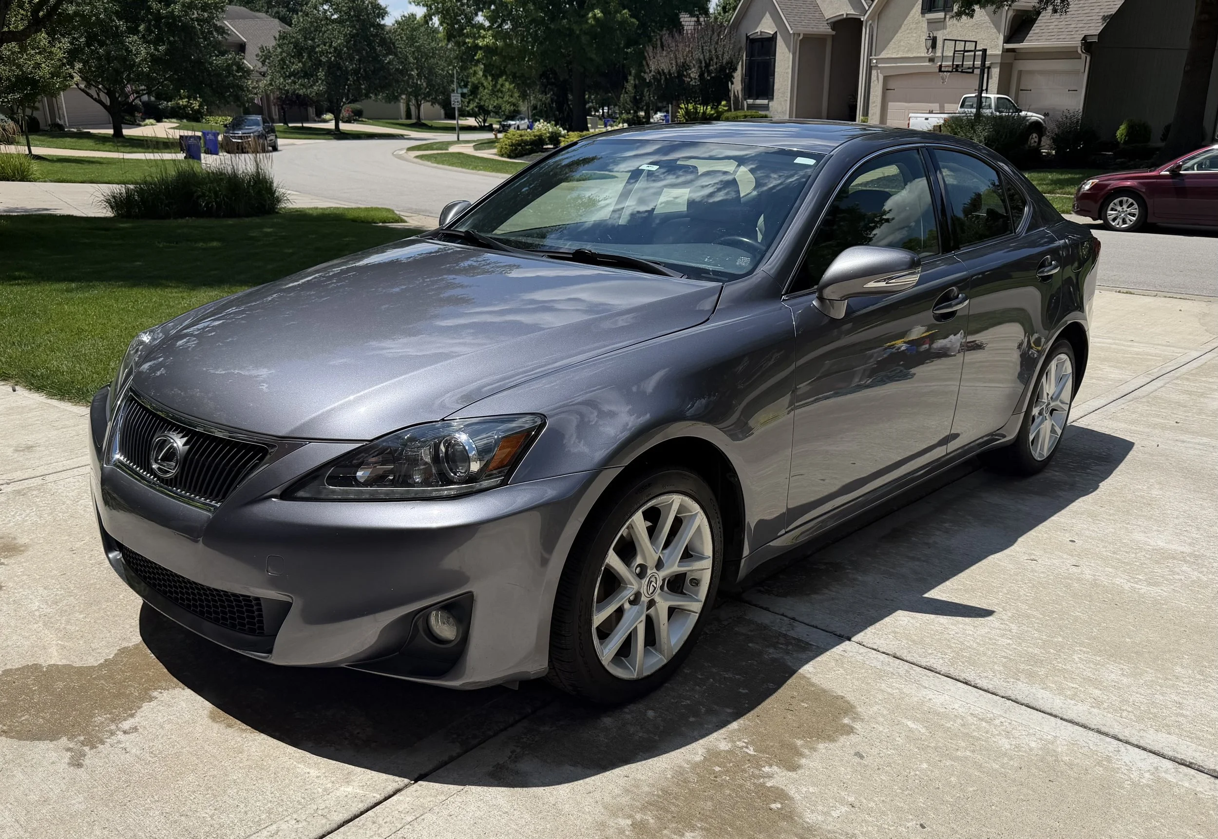 A silver Lexus sedan parked on a concrete driveway in a suburban neighborhood with houses, trees, and a street visible in the background.