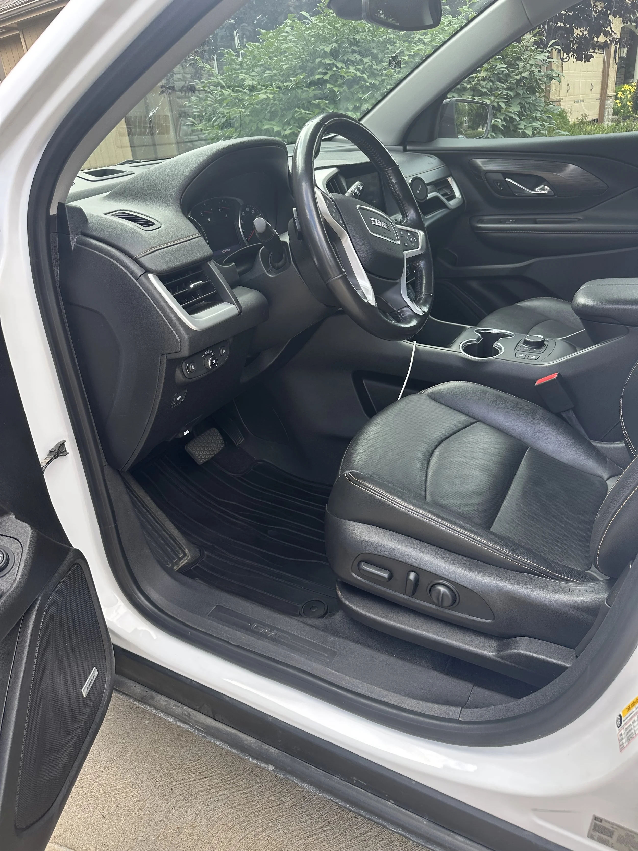 The interior of a GMC vehicle, showing the front passenger seat, dashboard, steering wheel, and center console with cup holders and controls. The door is open, revealing the exterior environment with greenery and a building in the background.