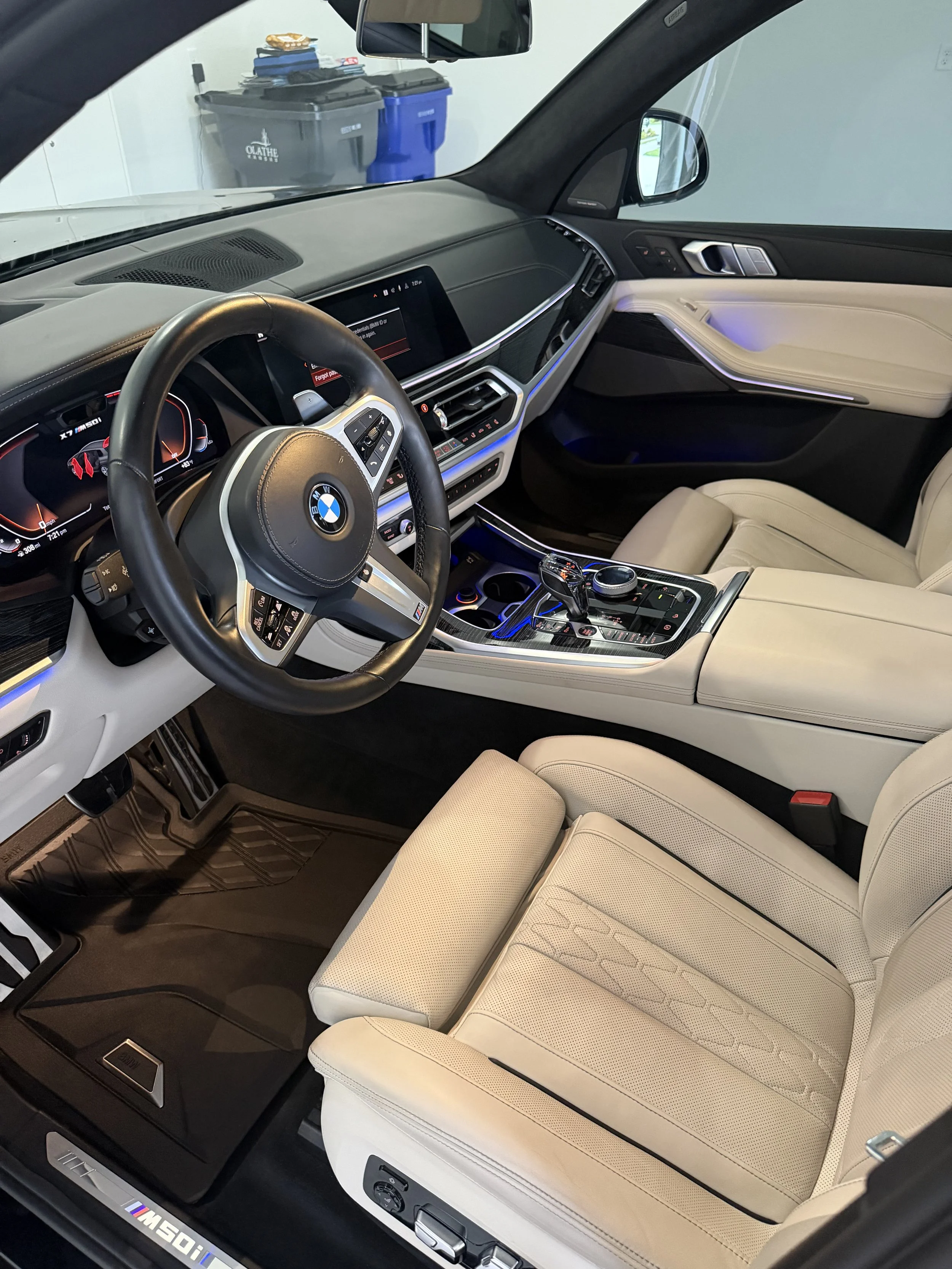 The interior of a BMW M50i SUV, featuring a black leather steering wheel with silver accents, a digital dashboard displaying various readings, a large center touchscreen, beige leather seats, and ambient lighting. In the background, two trash bins an