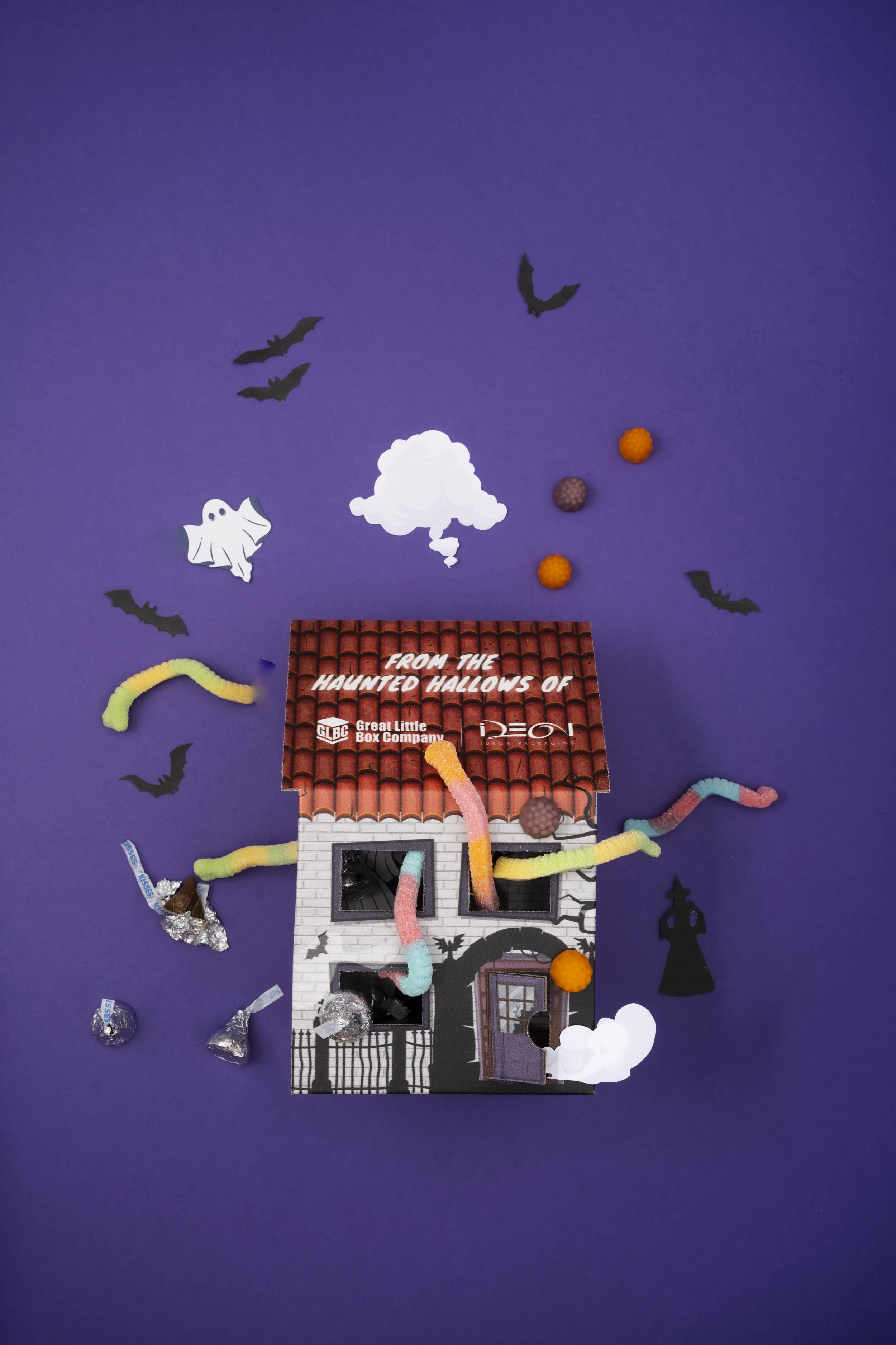 A Halloween-themed cardboard house decorated with paper and foam cutouts, gummy worms, candy, and black paper bats and ghosts on a purple background.