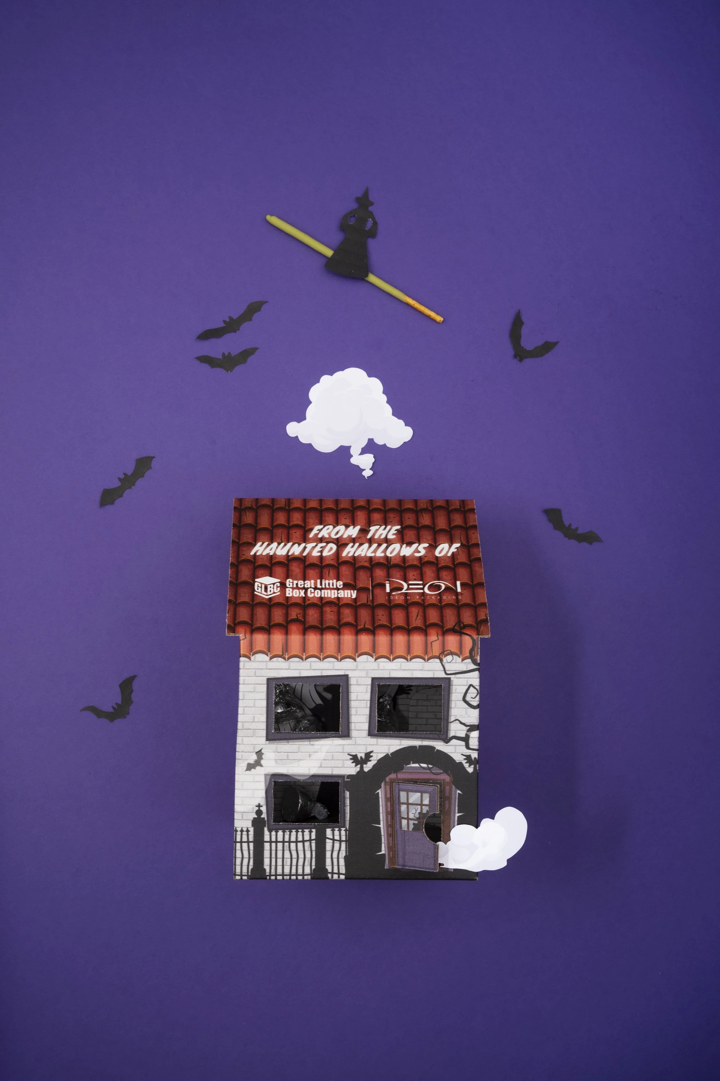 A handmade Halloween-themed paper house decoration with crow silhouettes, a witch silhouette on a clock, and a white cloud with a spelling mistake for 'hollows' on the roof, set against a purple background.