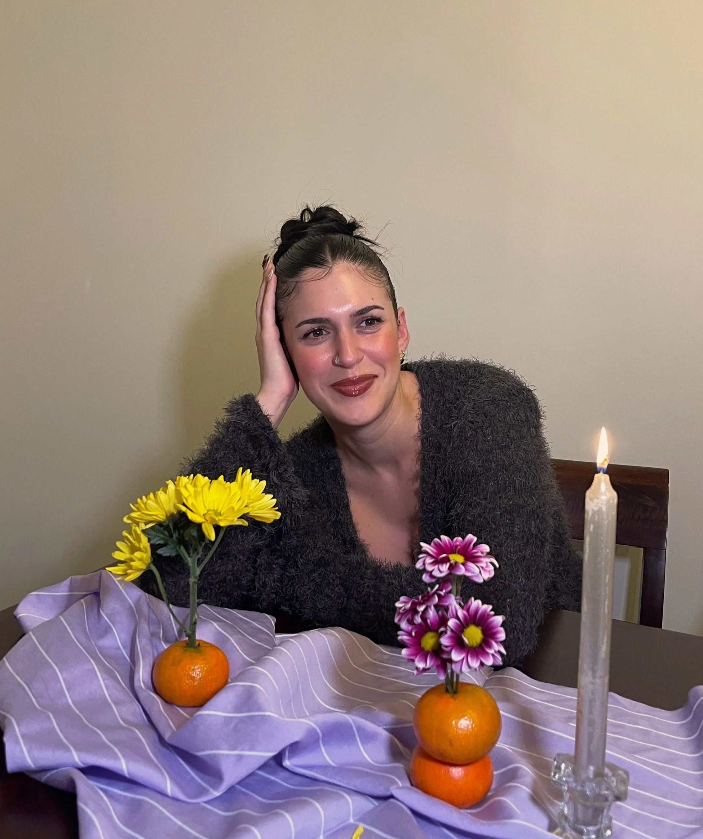 A woman with dark hair in a bun, wearing a black fuzzy sweater, is sitting at a table decorated with fruit and flowers, with a lit candle nearby. She is smiling and resting her head on her hand.