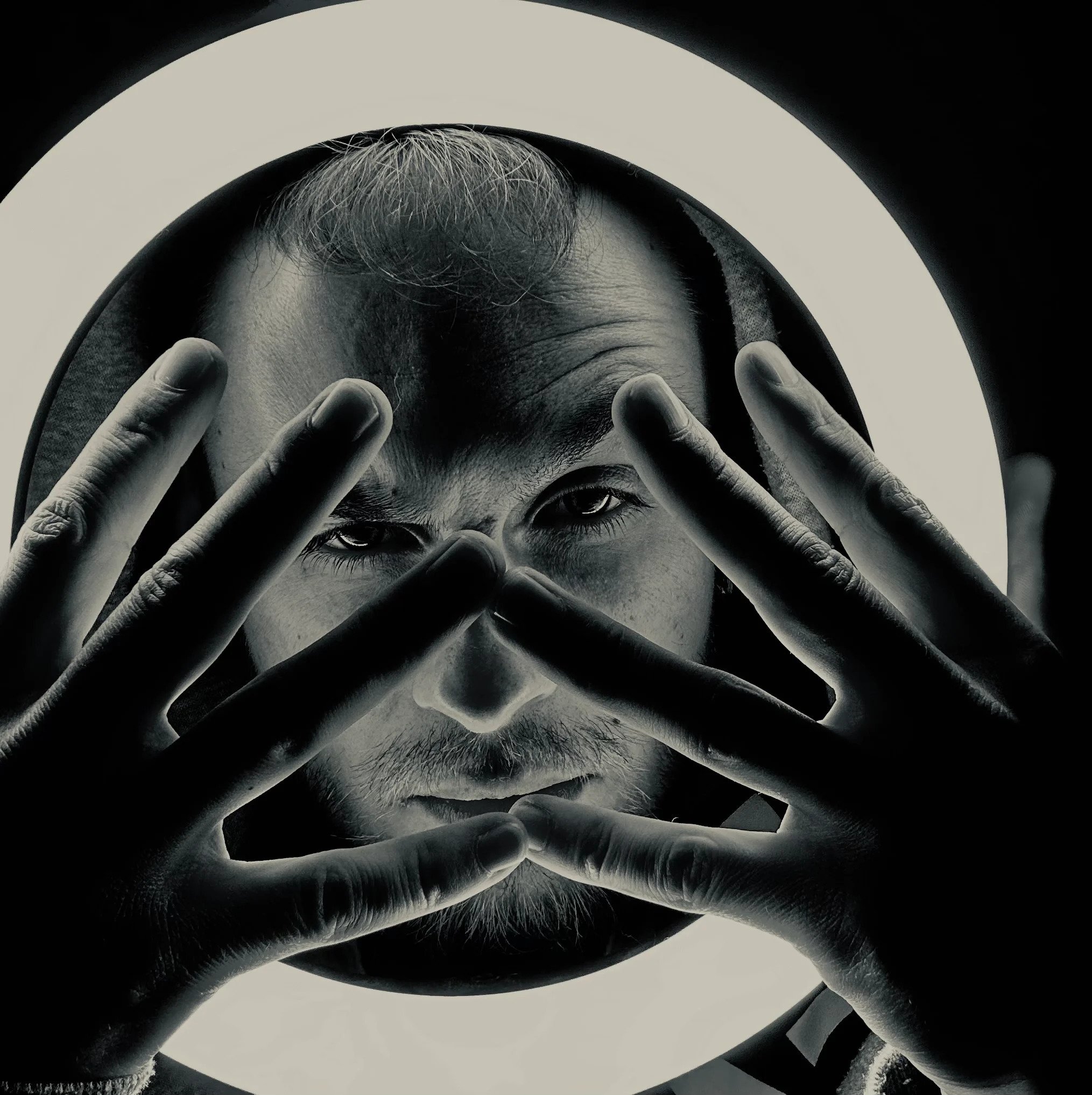 Schwarz-weiß Porträt eines Mannes mit Bart, der durch seine Hände schaut, die vor seinem Gesicht gehalten sind. Ein ringförmiges Licht umrahmt sein Gesicht im Hintergrund.