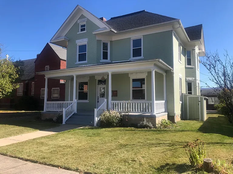A two-story light green house with a large front porch and white railing, black roof, surrounded by a lawn with a sidewalk leading to the front steps, and other houses visible in the background.