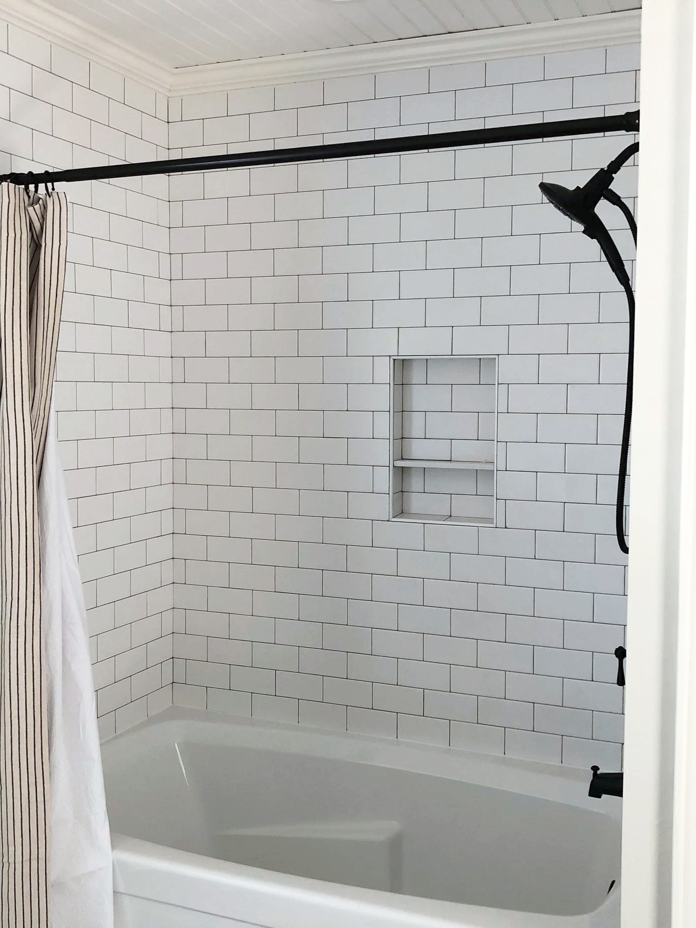 A clean bathtub and shower with white subway tile walls, a niche in the wall, a mounted showerhead, and a black curtain rod with a striped curtain.