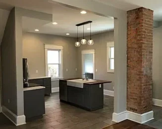 Modern kitchen with island, pendant lighting, exposed brick chimney, warm gray walls, and three windows.