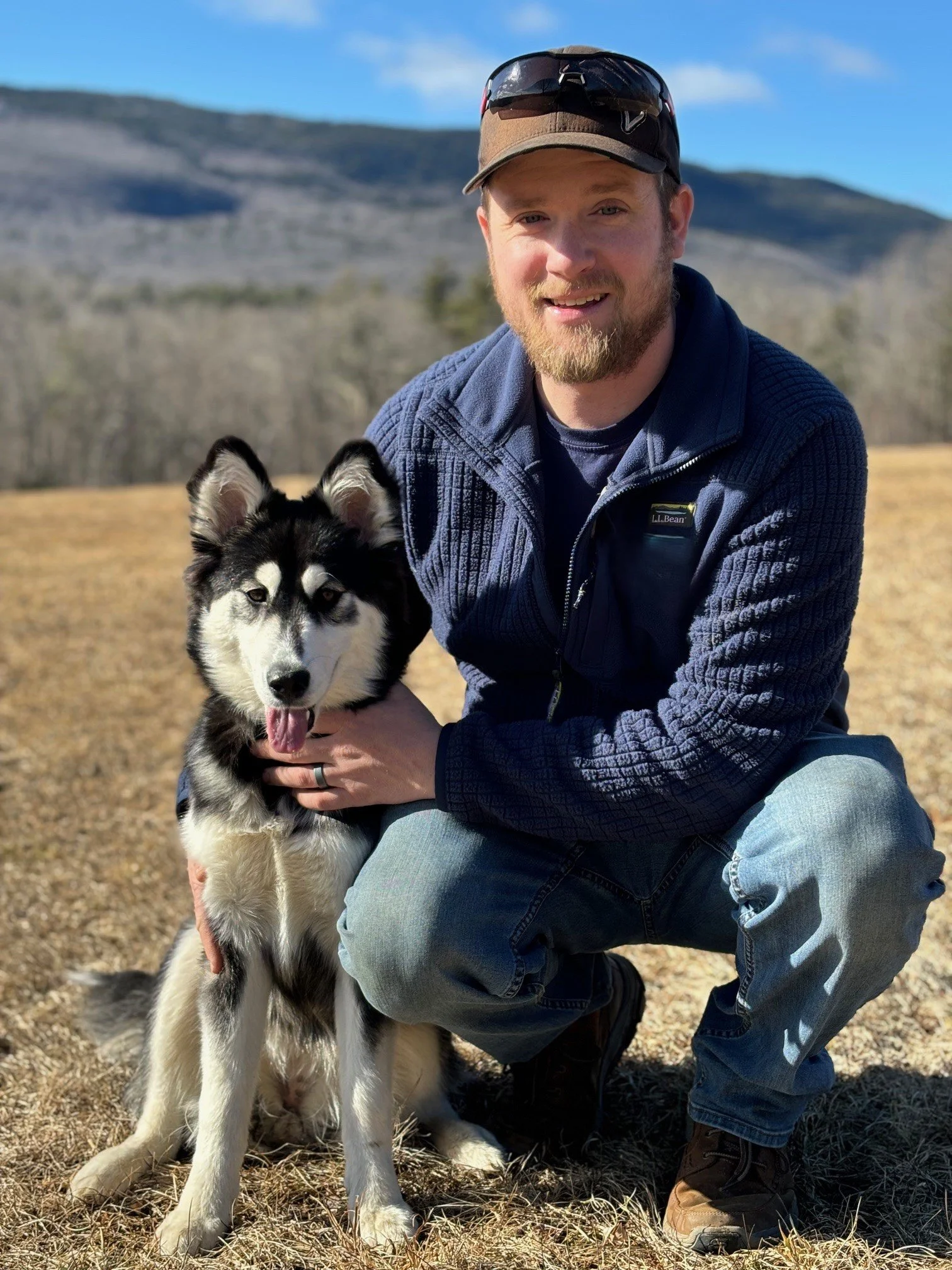 A man in outdoor clothing squats next to a black and white Siberian Husky dog in a grassy field with mountains and trees in the background.