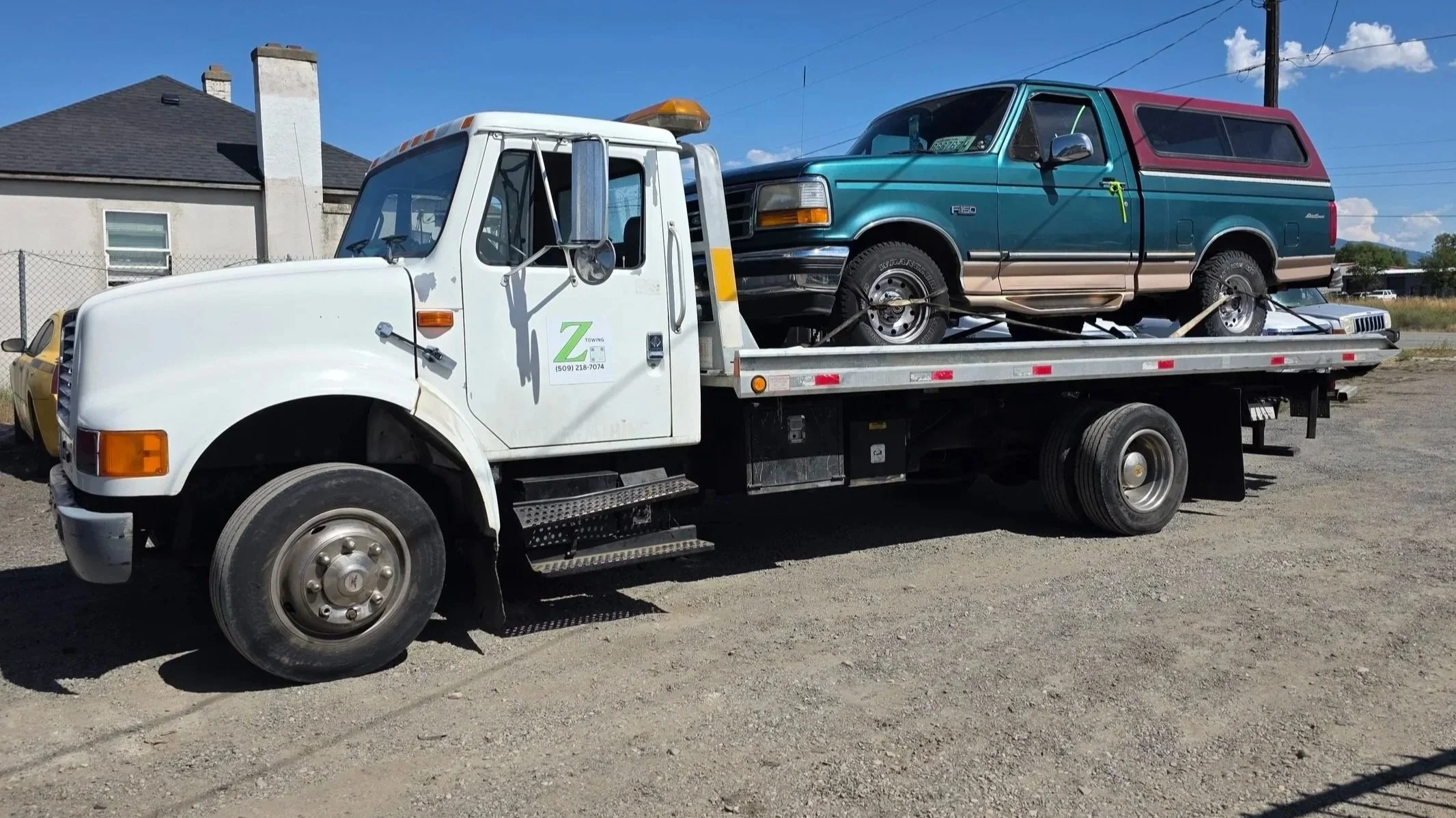 A white flatbed tow truck carrying a teal and maroon Chevrolet Astro van on its bed, parked on a dirt lot with a house and a blue sky in the background.