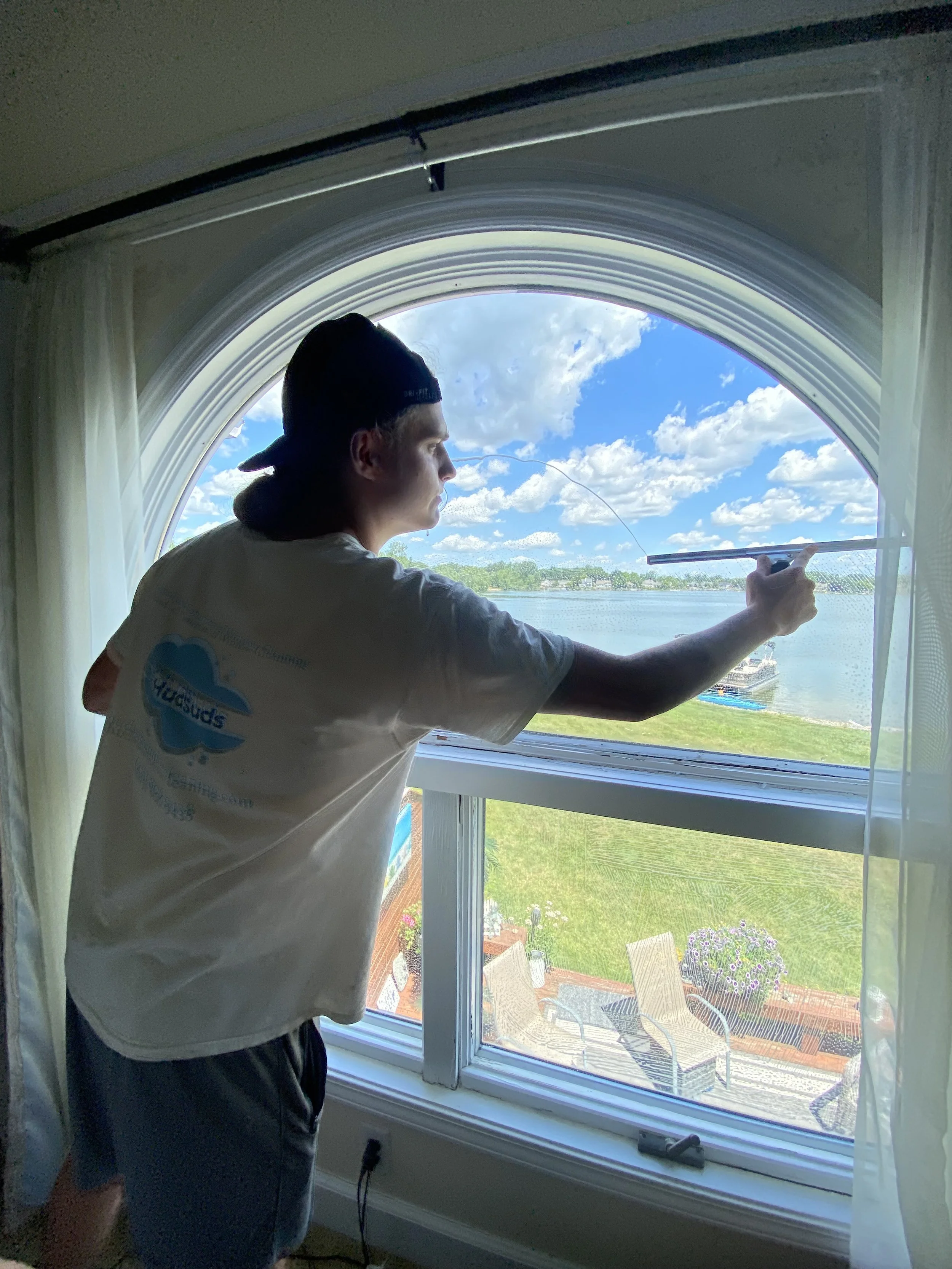 A young man in a baseball cap and t-shirt cleaning the inside of a large arched window with a squeegee, overlooking a lakeside with chairs, a flowerpot with purple flowers, and a blue sky with white clouds.