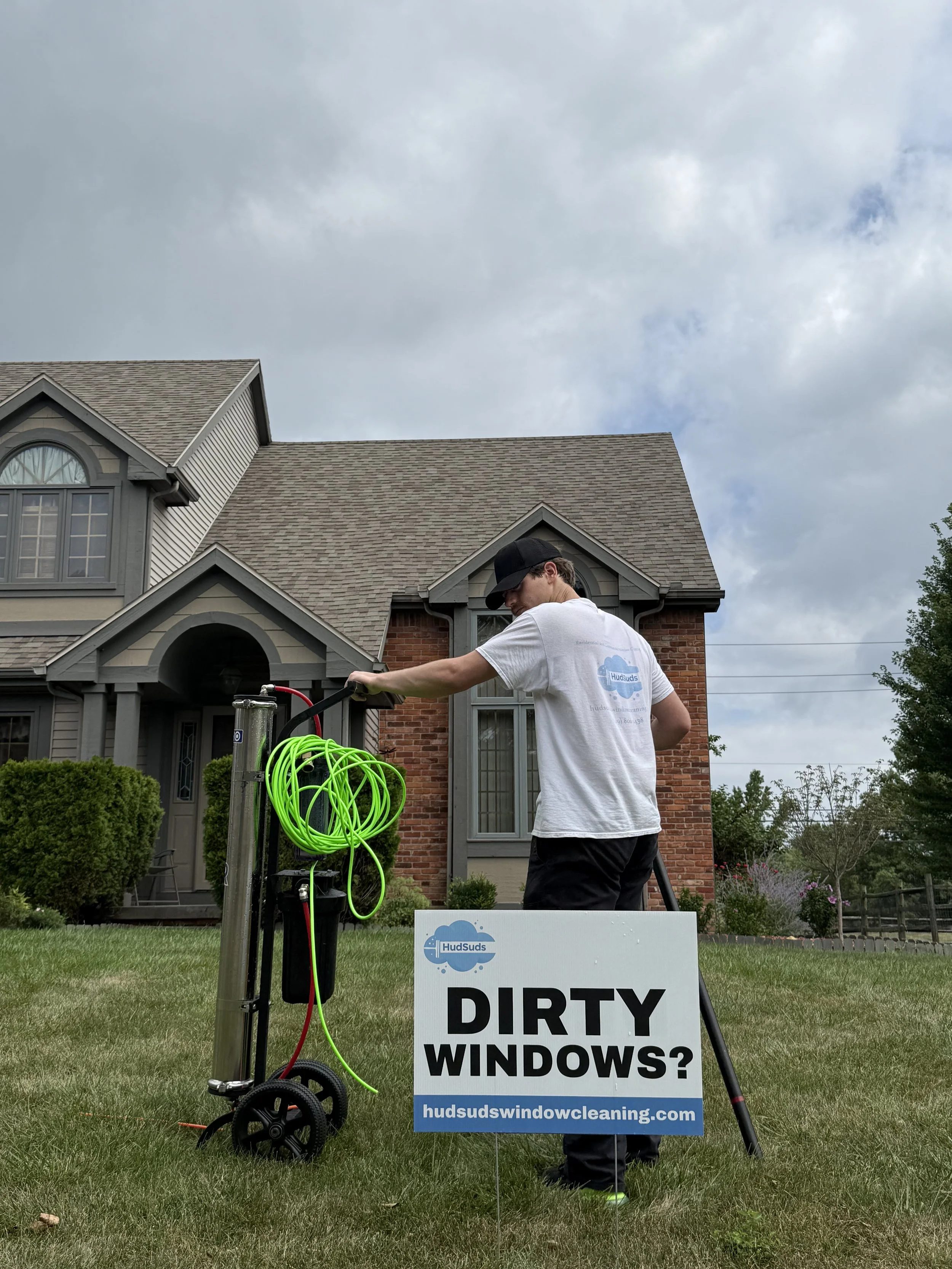 A window cleaner working outside a house with a sign that reads "Dirty Windows? hudsudswindowcleaning.com". The cleaner is adjusting equipment on a cart, wearing a white t-shirt and a black cap, with a cloudy sky overhead.
