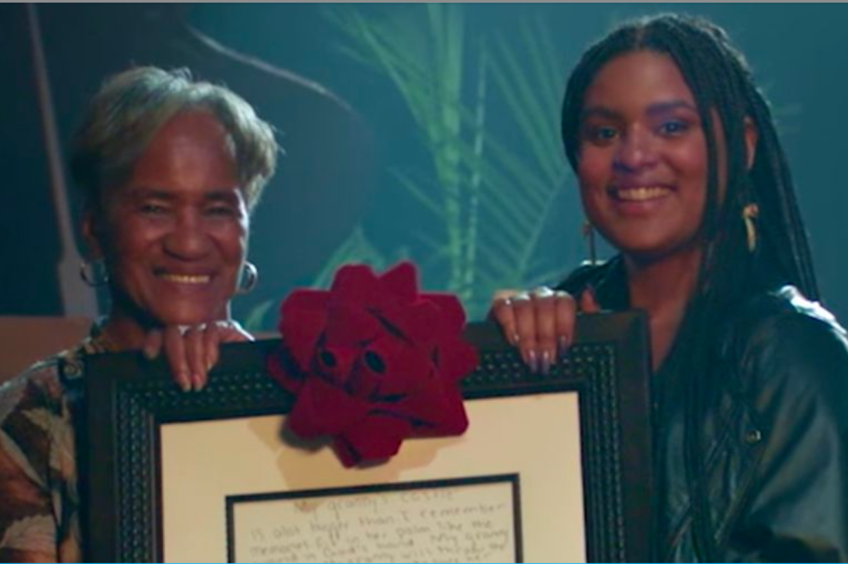 Two women smiling and holding a framed certificate decorated with a large red flower, with a background of green plants.