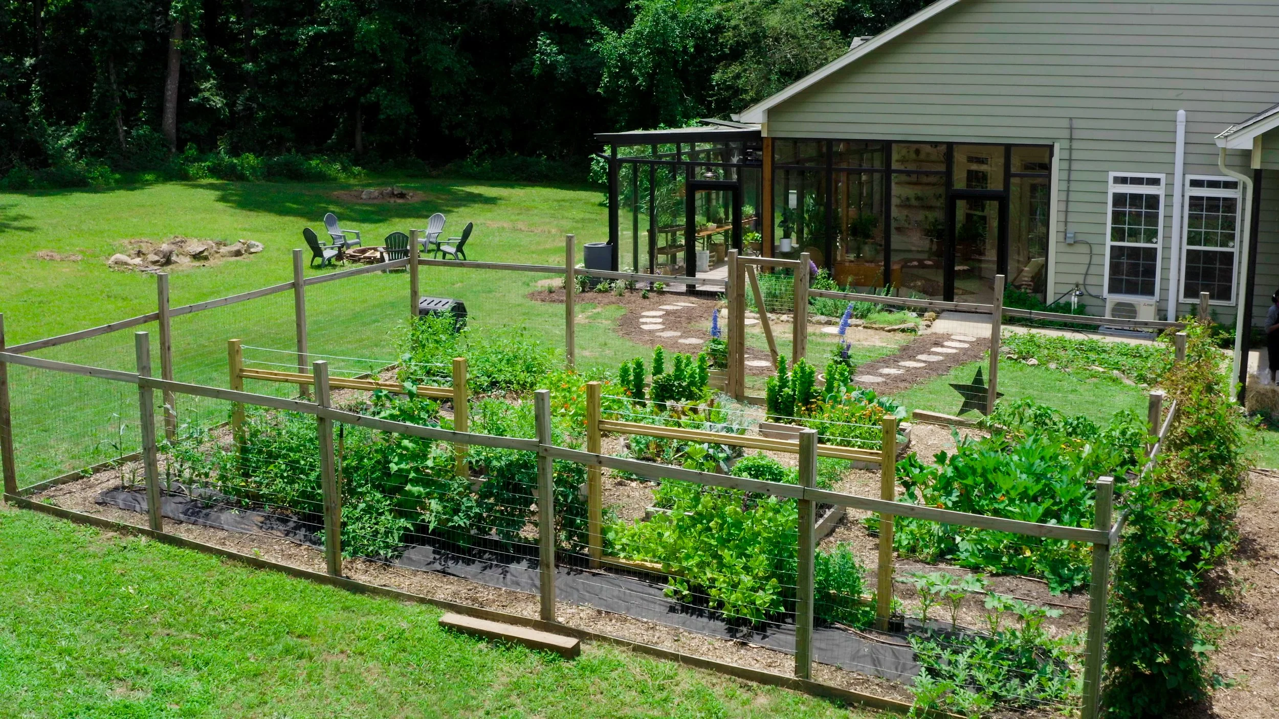 A backyard vegetable garden enclosed by a wooden fence, with a house featuring a glass sunroom in the background, a walking stone path, and an outdoor seating area with four black chairs and a fire pit.