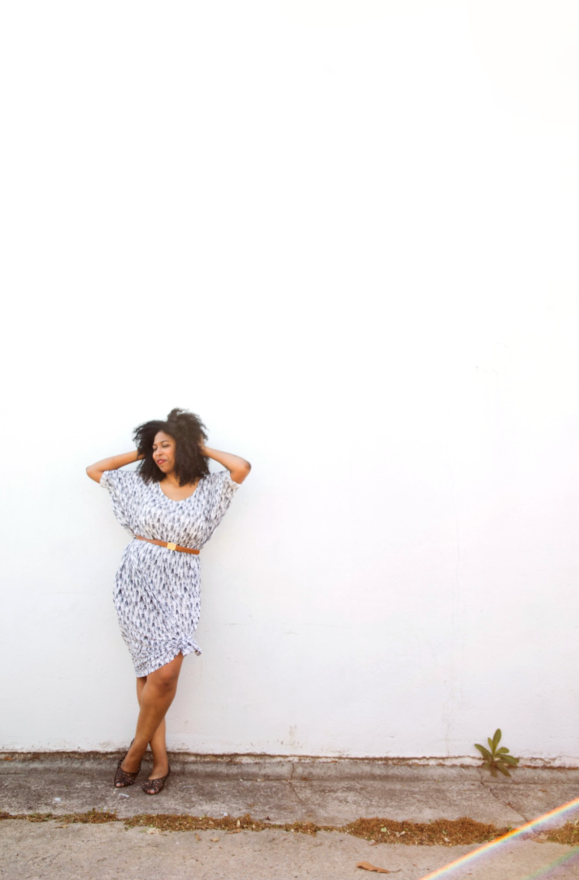 Lesly Washigngton standing against a white wall with her eyes closed, smiling, wearing a patterned dress, with her hands behind her head, and a small green plant near her feet.