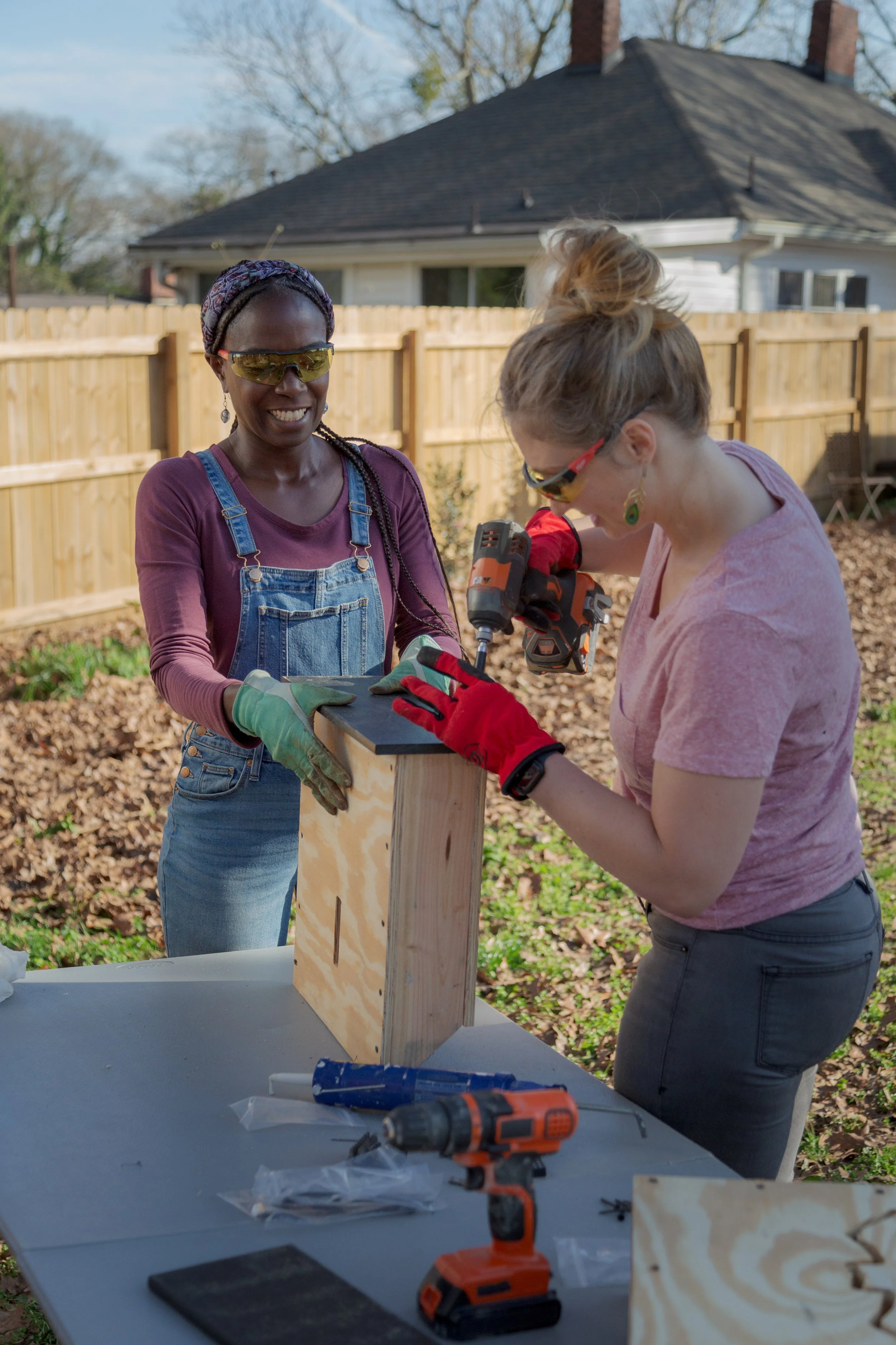 Two women working on a woodworking project outdoors, one holding a piece of wood steady while the other screws it into place with a power drill. They are wearing gloves and protective eyewear, with tools and wood on a table in front of them.