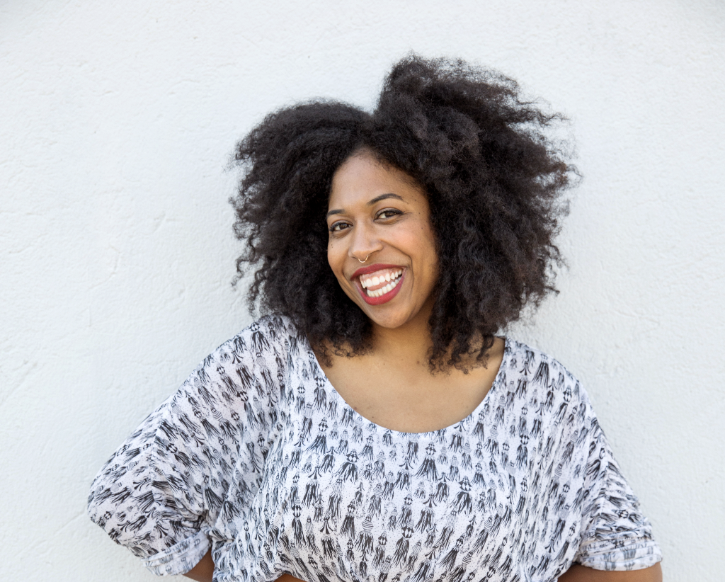 Lesly Washington, a woman with curly black hair smiling and standing against a plain white wall.