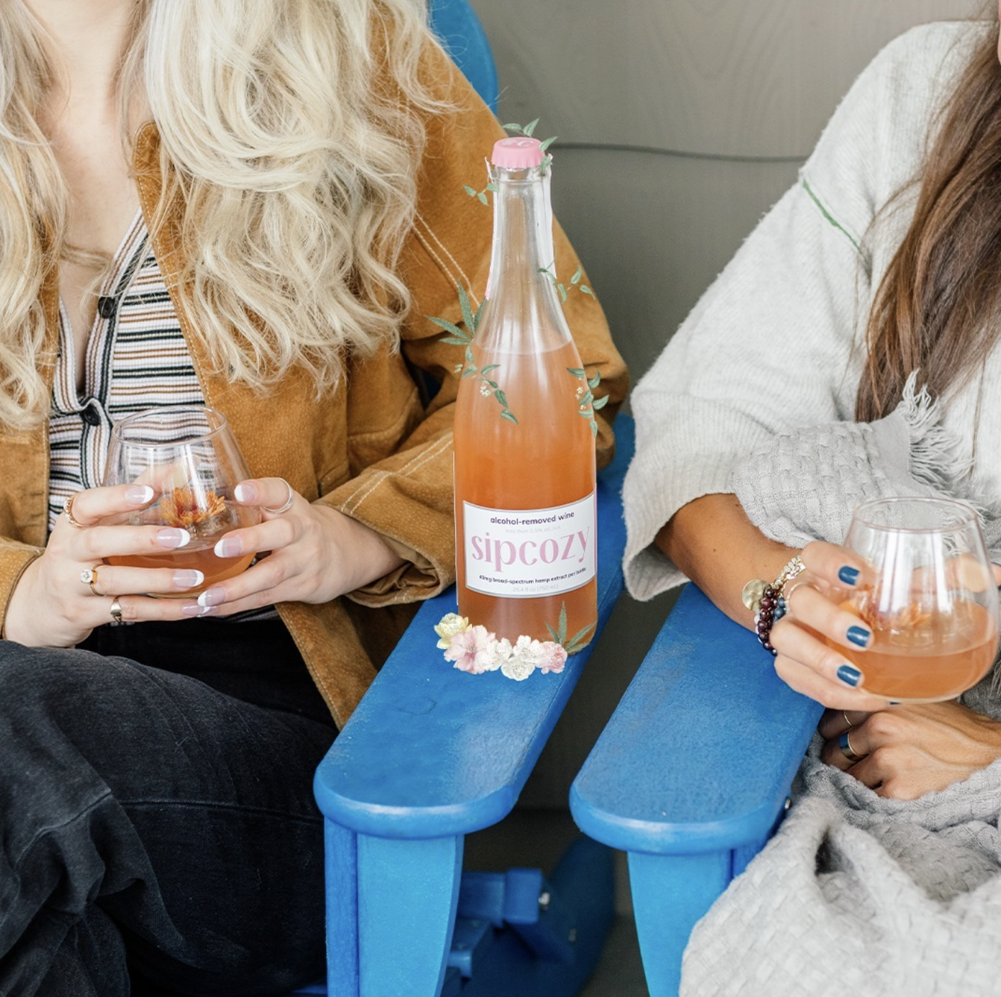 Two women sitting at a blue table, each holding a glass of pink-colored beverage, with a bottle labeled 'sipcozy' on the table between them.
