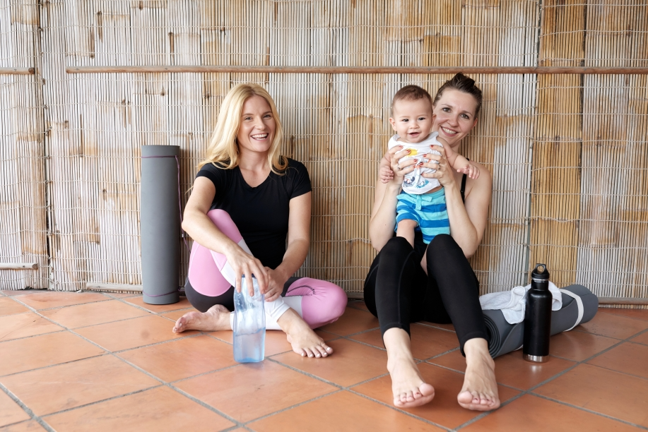 Two women and a baby sitting on a tiled floor with yoga mats; one woman is holding a water bottle, the other is holding the baby, all smiling.