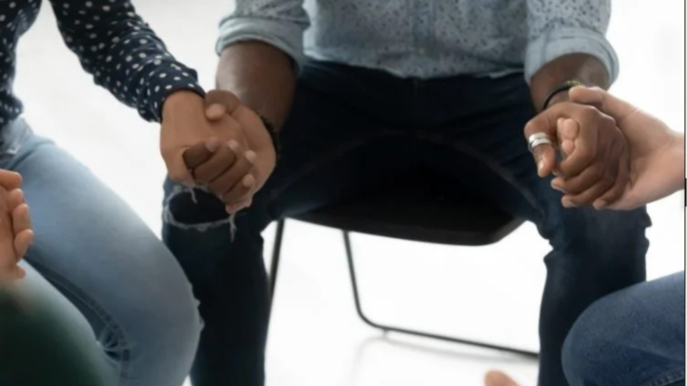 A group of people sitting and holding hands in a circle, possibly during a support meeting or group therapy.