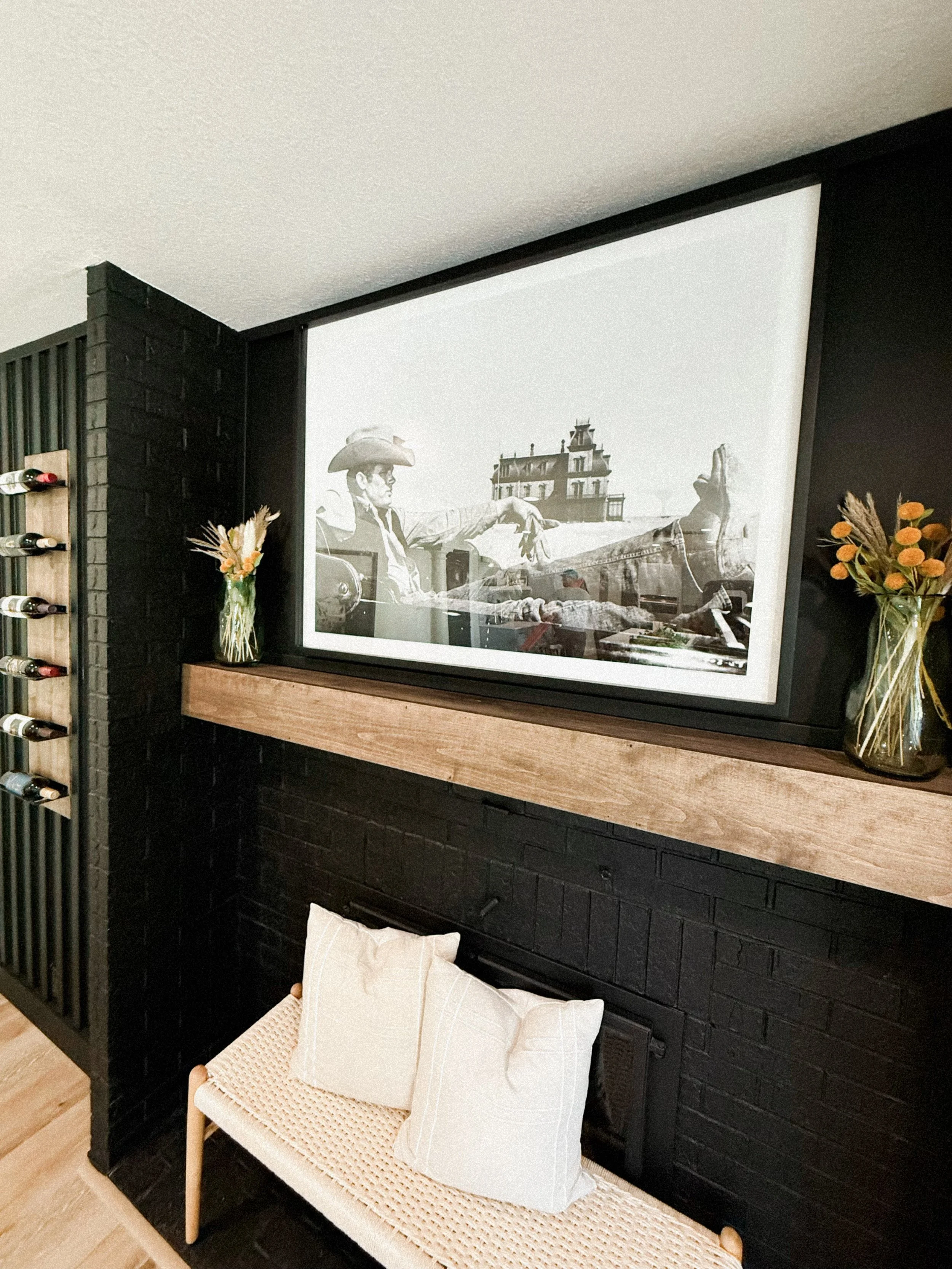 A room with a black brick wall, a wooden shelf above a fireplace, large flat-screen TV displaying a black and white image of a cowboy, two small vases with dried flowers on either side of the TV, and a small woven bench with two white pillows in fron