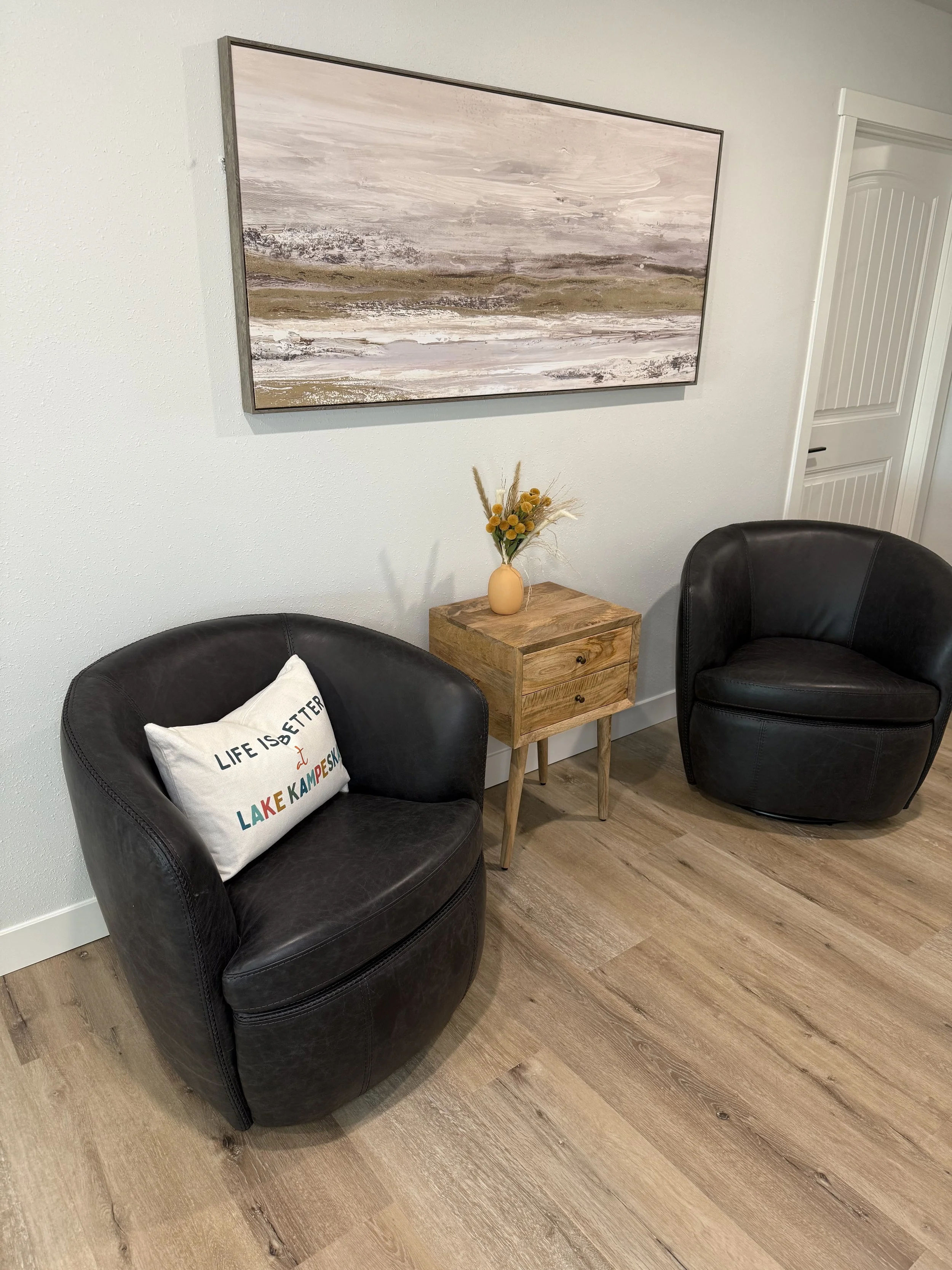 A cozy sitting area with two black leather armchairs, a small wooden side table, a decorative pillow with the phrase "Life is better at Lake Kampeska," a vase with yellow flowers and grasses, a landscape painting on the wall, and hardwood flooring.