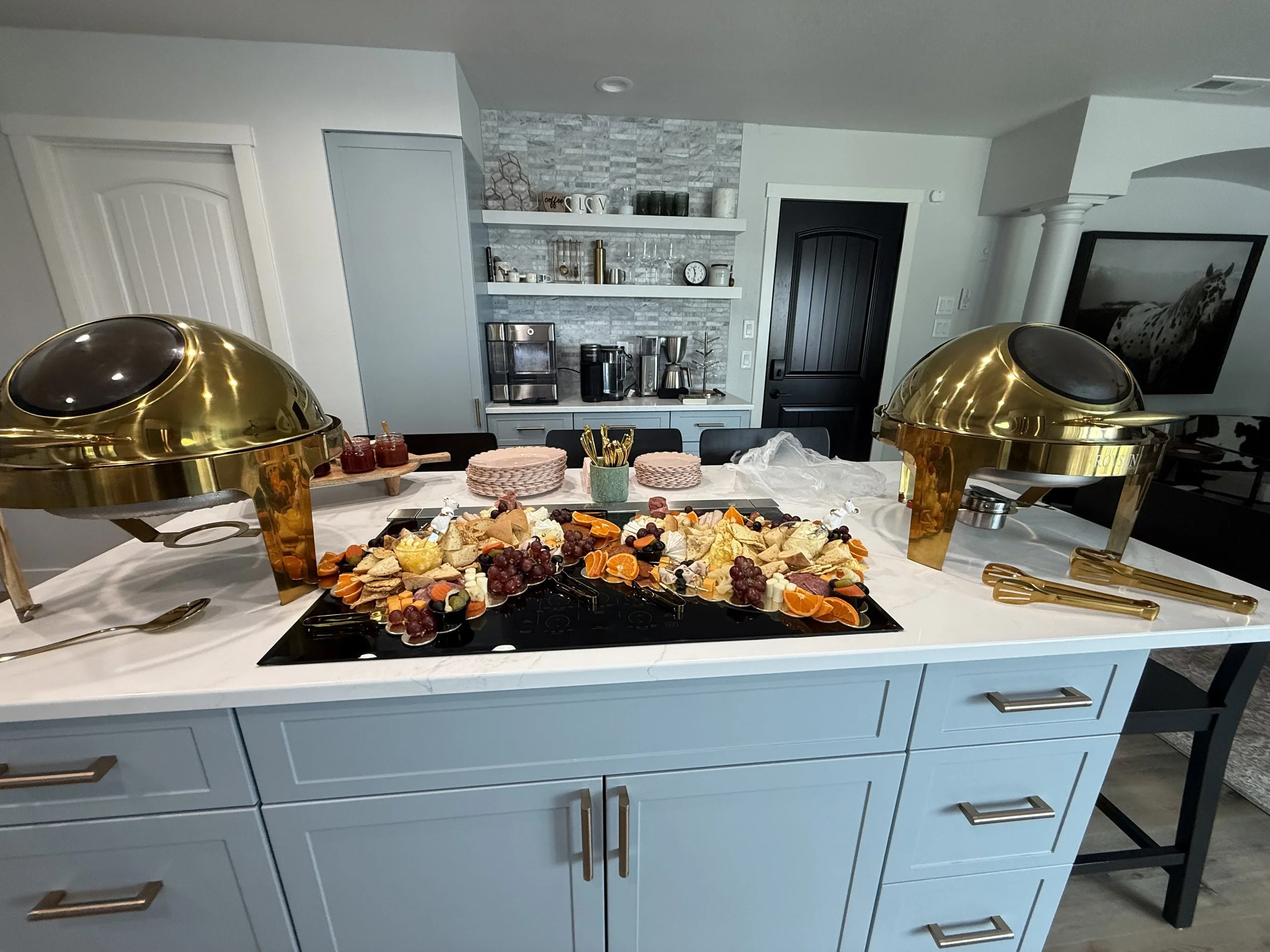Buffet table with cheese, crackers, grapes, oranges, and other fruits, with chafing dishes and serving utensils in a modern kitchen.